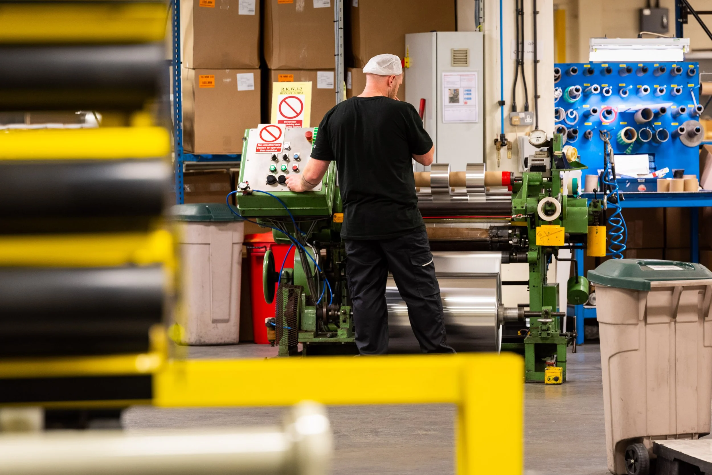 Worker controlling foil roll processing equipment inside a busy industrial warehouse.