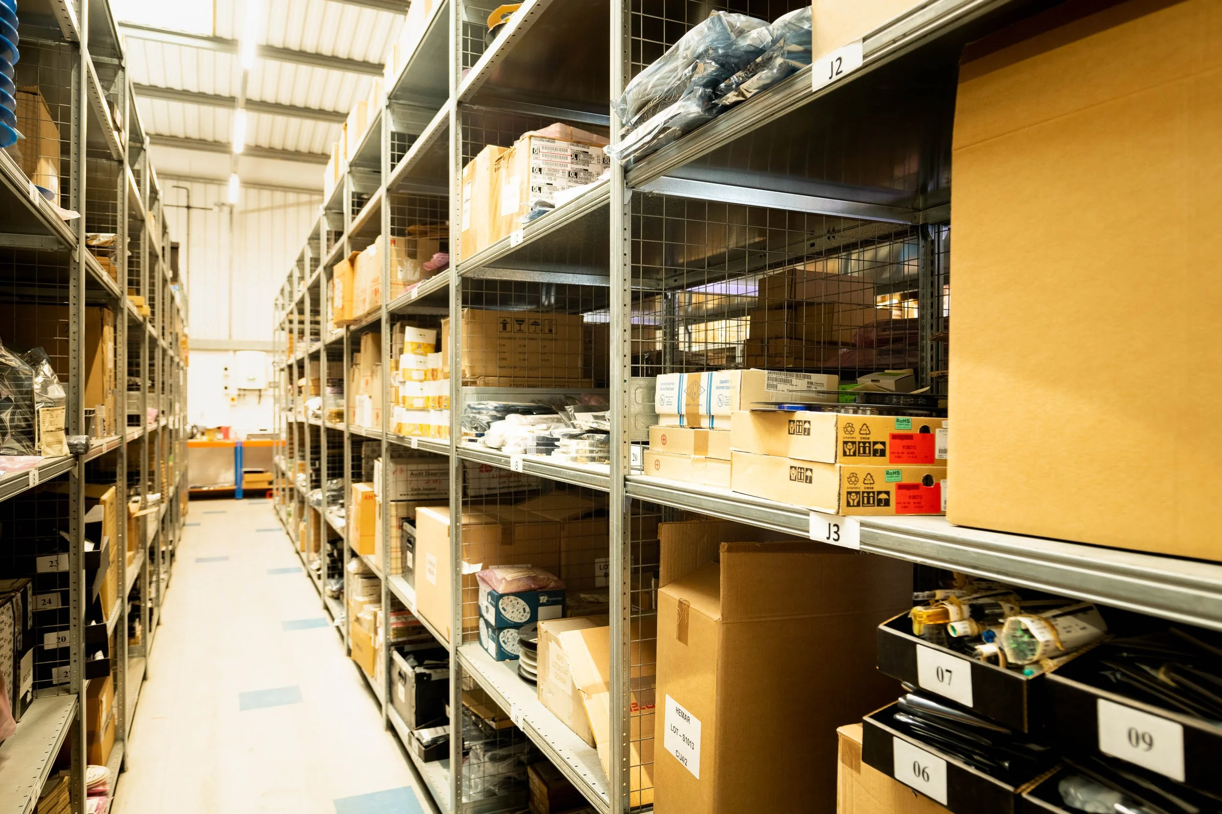 Long aisle of industrial warehouse with labelled storage shelves stacked with boxes and equipment.