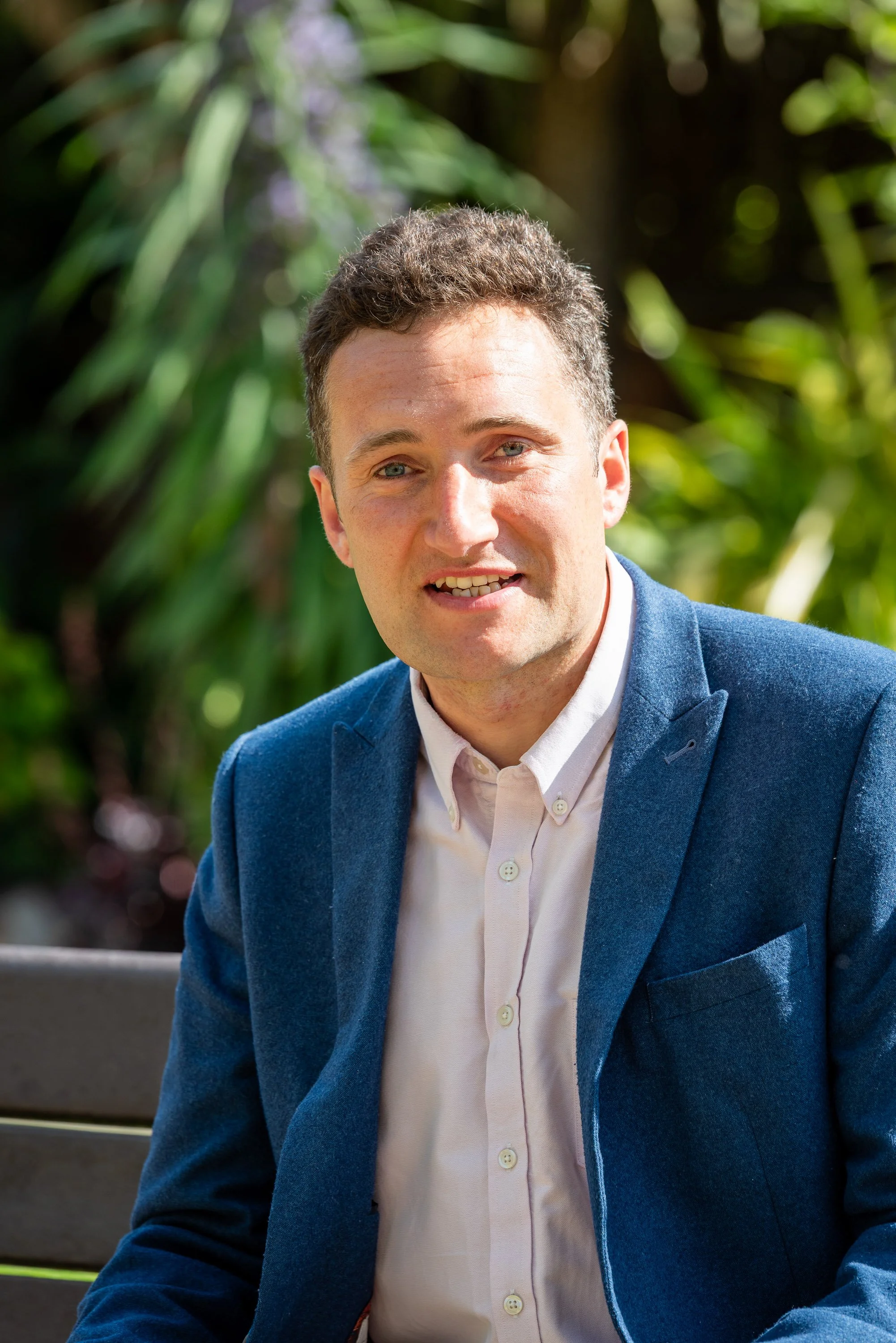 Outdoor professional headshot of a man in a blazer with a natural blurred background.