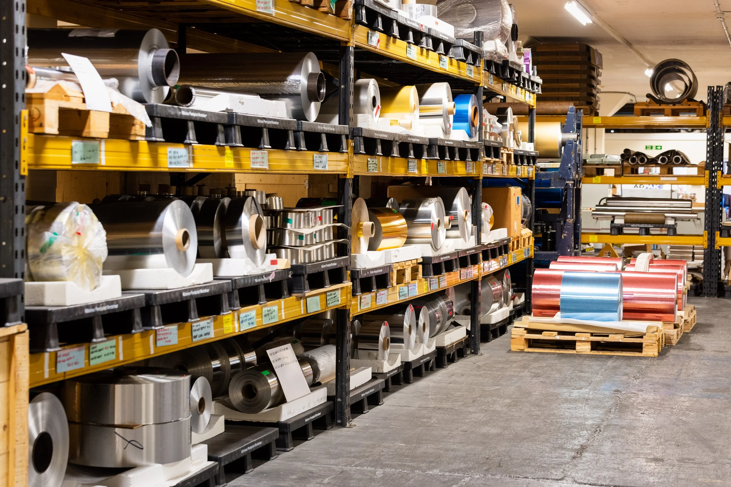 Industrial warehouse storage racks filled with aluminium foil rolls in various finishes and colours.