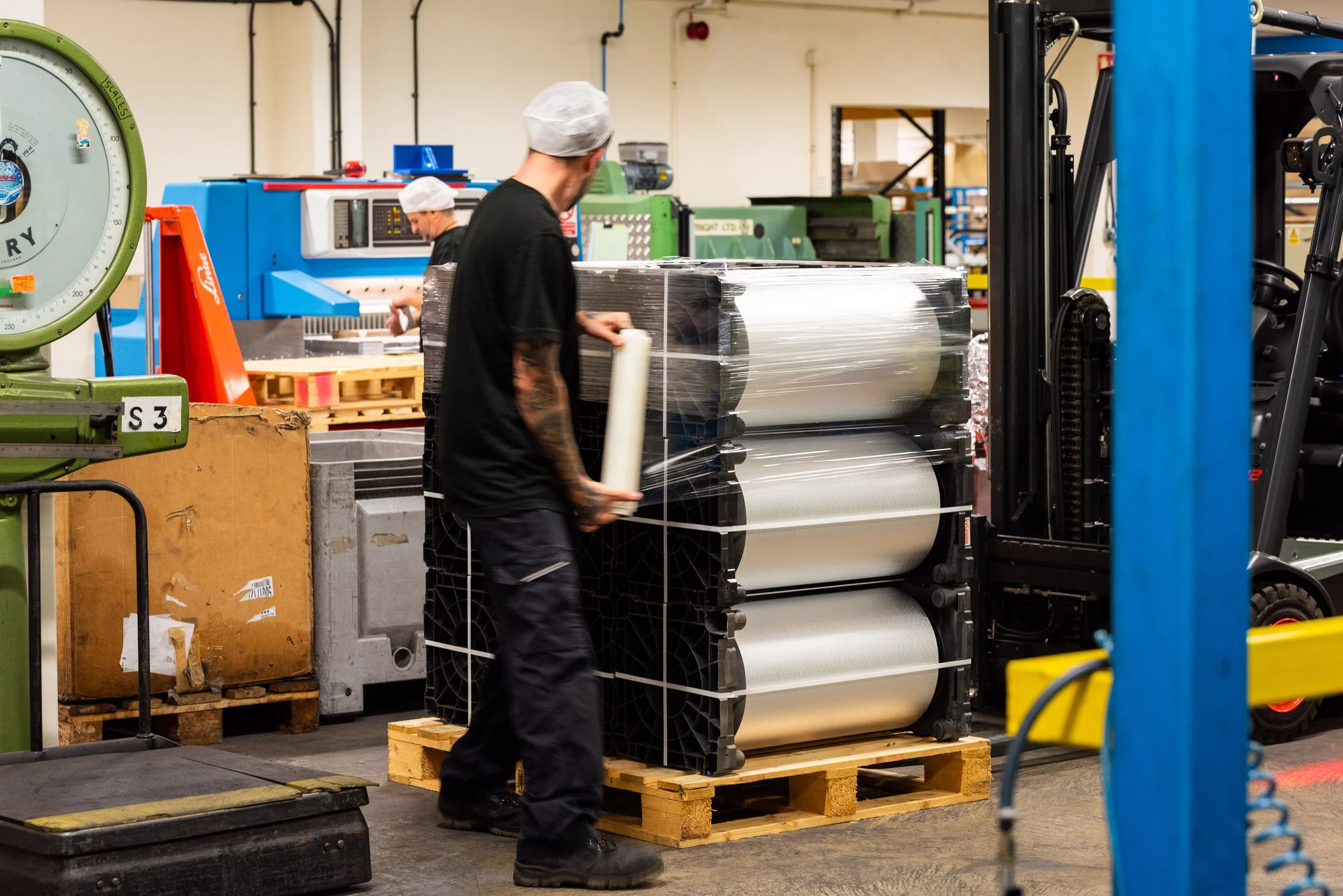 Worker wrapping aluminium foil rolls on pallet in manufacturing warehouse with industrial equipment.