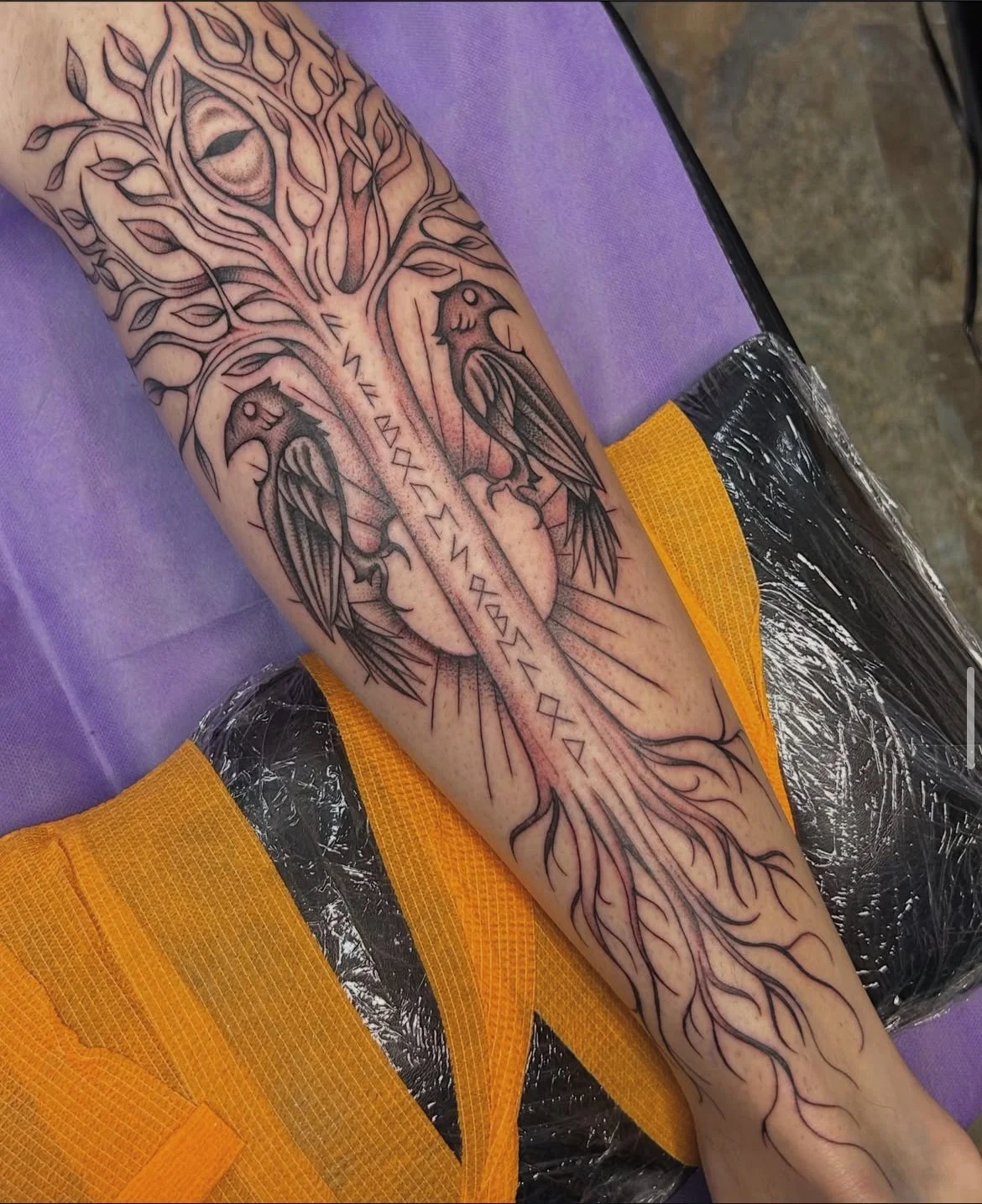A tattoo of a tree with birds, runes, and intricate roots on an arm, in progress at a tattoo shop.