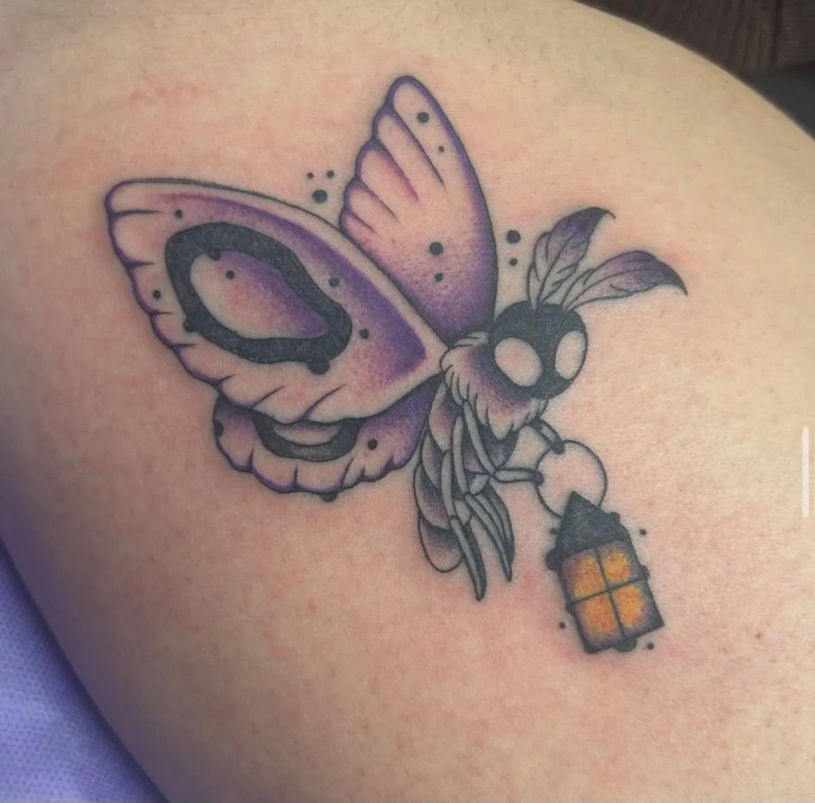 A colorful tattoo of a butterfly with purple and black wings, a skull with feathered eyes, and a small lantern on light skin.