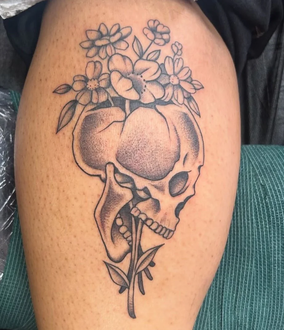 Tattoo of a skull with flowers and leaves growing out of it on a person's thigh.