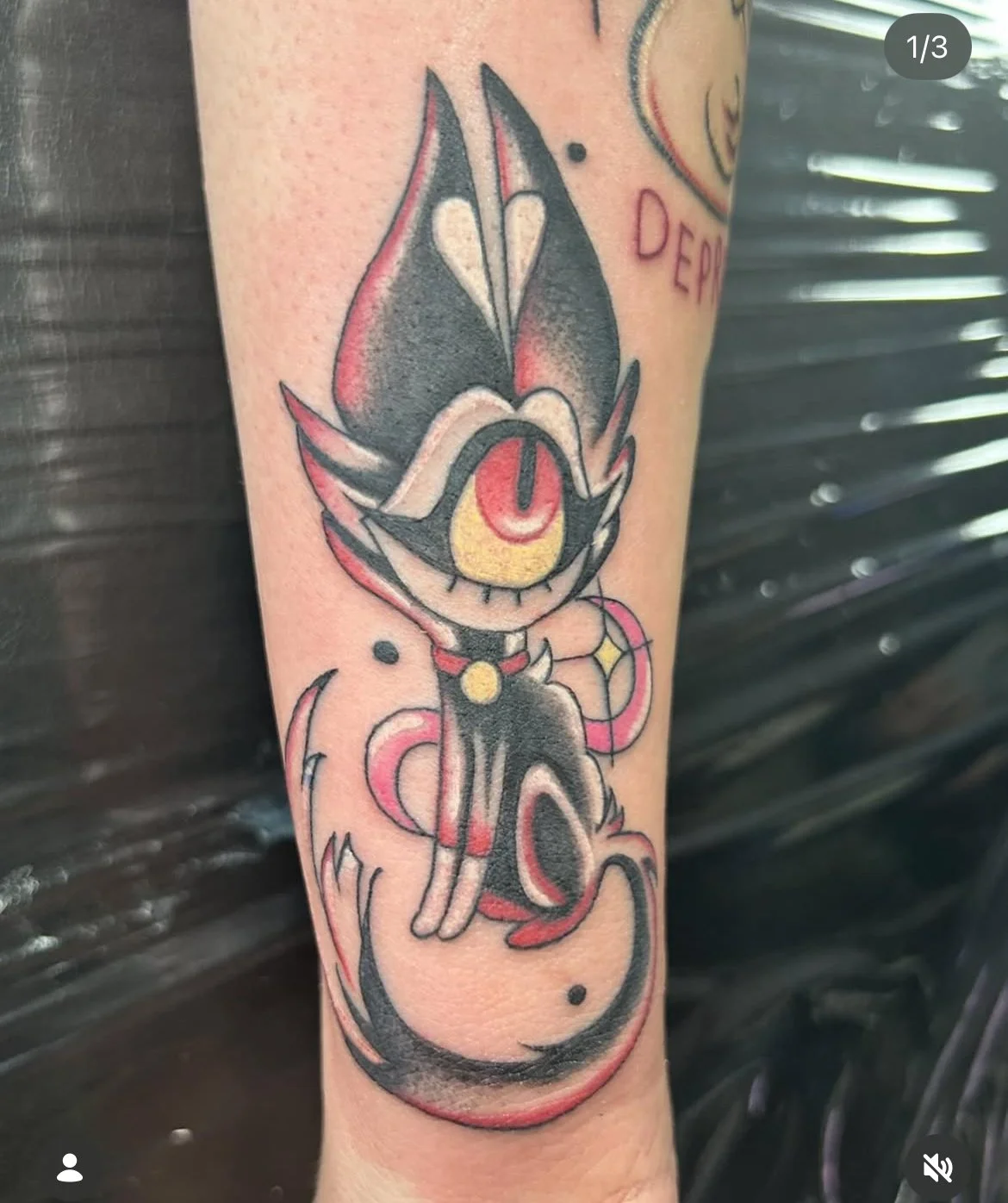 Tattoo of a cartoon-style black cat with a large yellow eye and a red slit pupil, sitting with a mischievous expression, surrounded by pink and black swirls. hazbin hotel