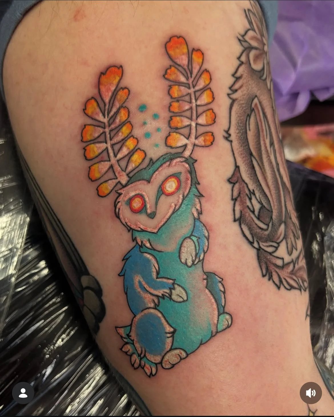 Colorful tattoo of a cute, chubby creature with large red eyes, sitting and holding one paw up, with orange and yellow leaf-like antlers on its head, and blue and pink fur.