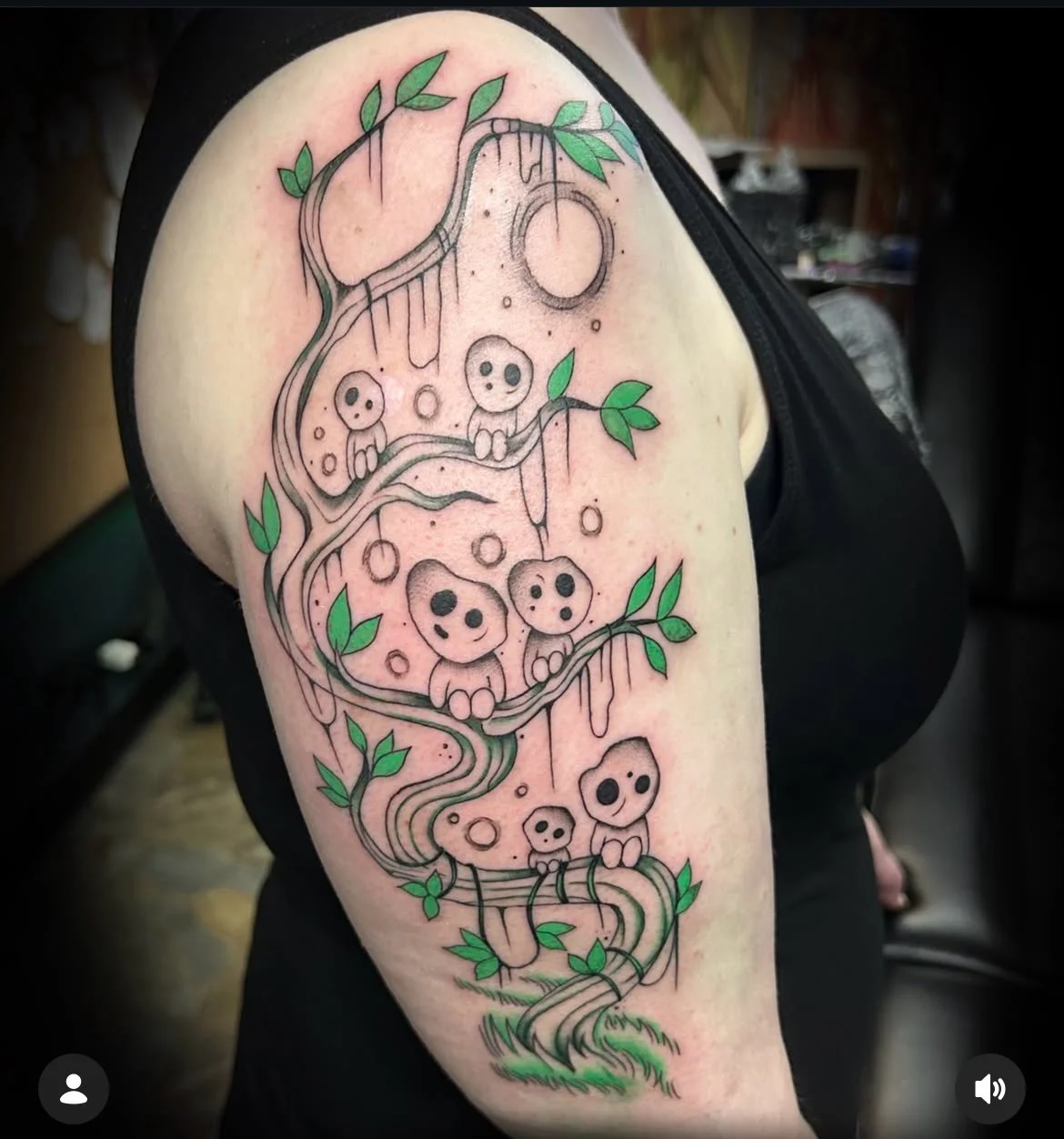 Tattoo of a whimsical, thin-lined black and green design featuring mushroom-like creatures with black eyes, a winding vine with green leaves, and dripping elements on the person's upper arm.