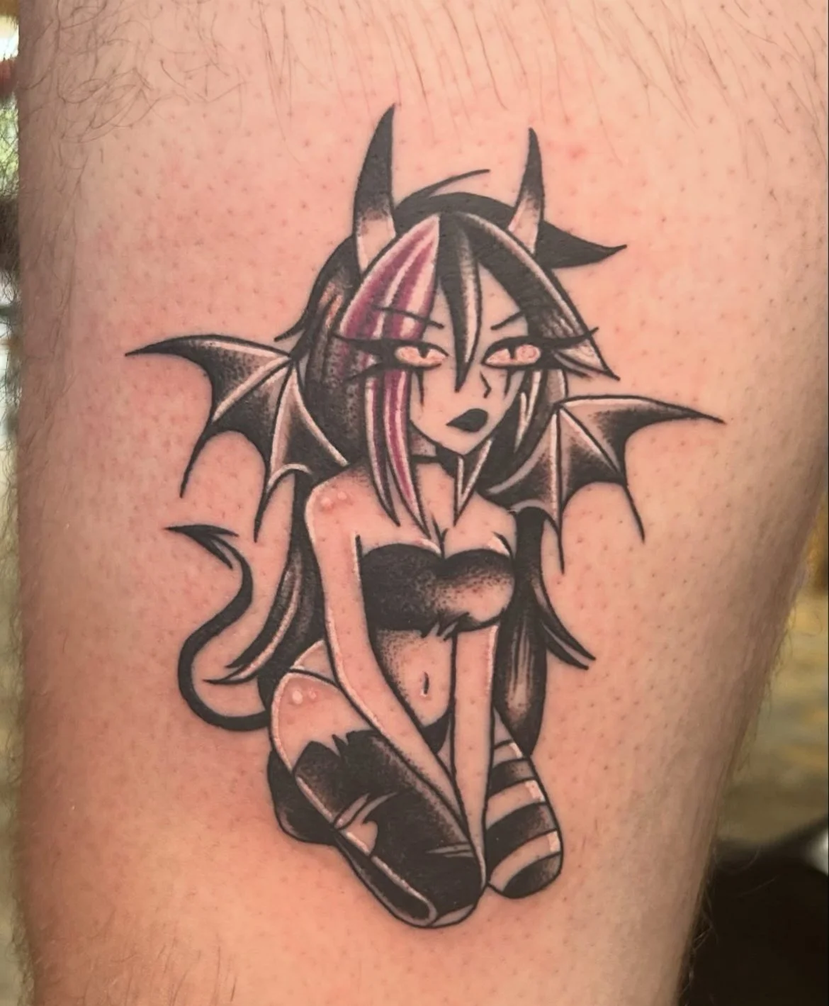 Tattoo of a fantasy girl with horns, bat-like wings, and striped socks, sitting with her knees pulled up, in black and pink ink on skin.