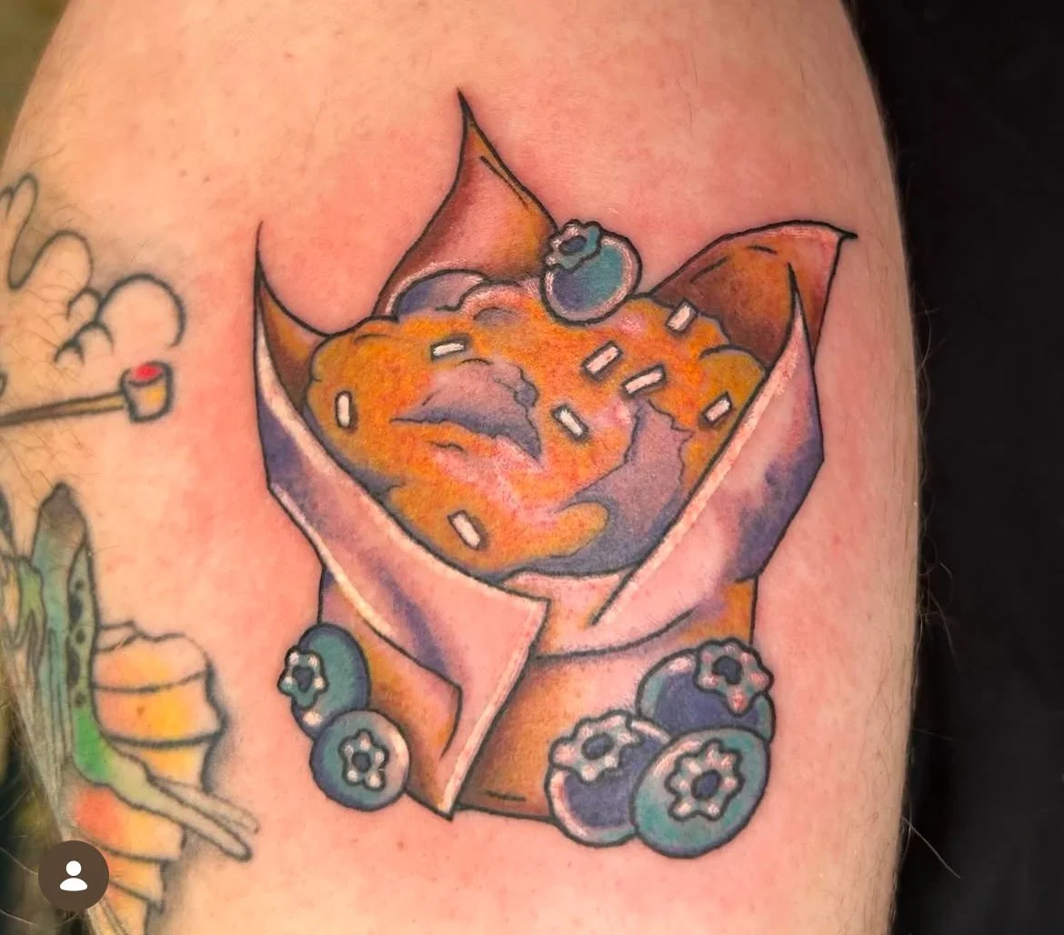 A colorful tattoo of a slice of pie with blueberries on top and small white rectangles resembling sprinkles or sugar, surrounded by blueberries and flowers.