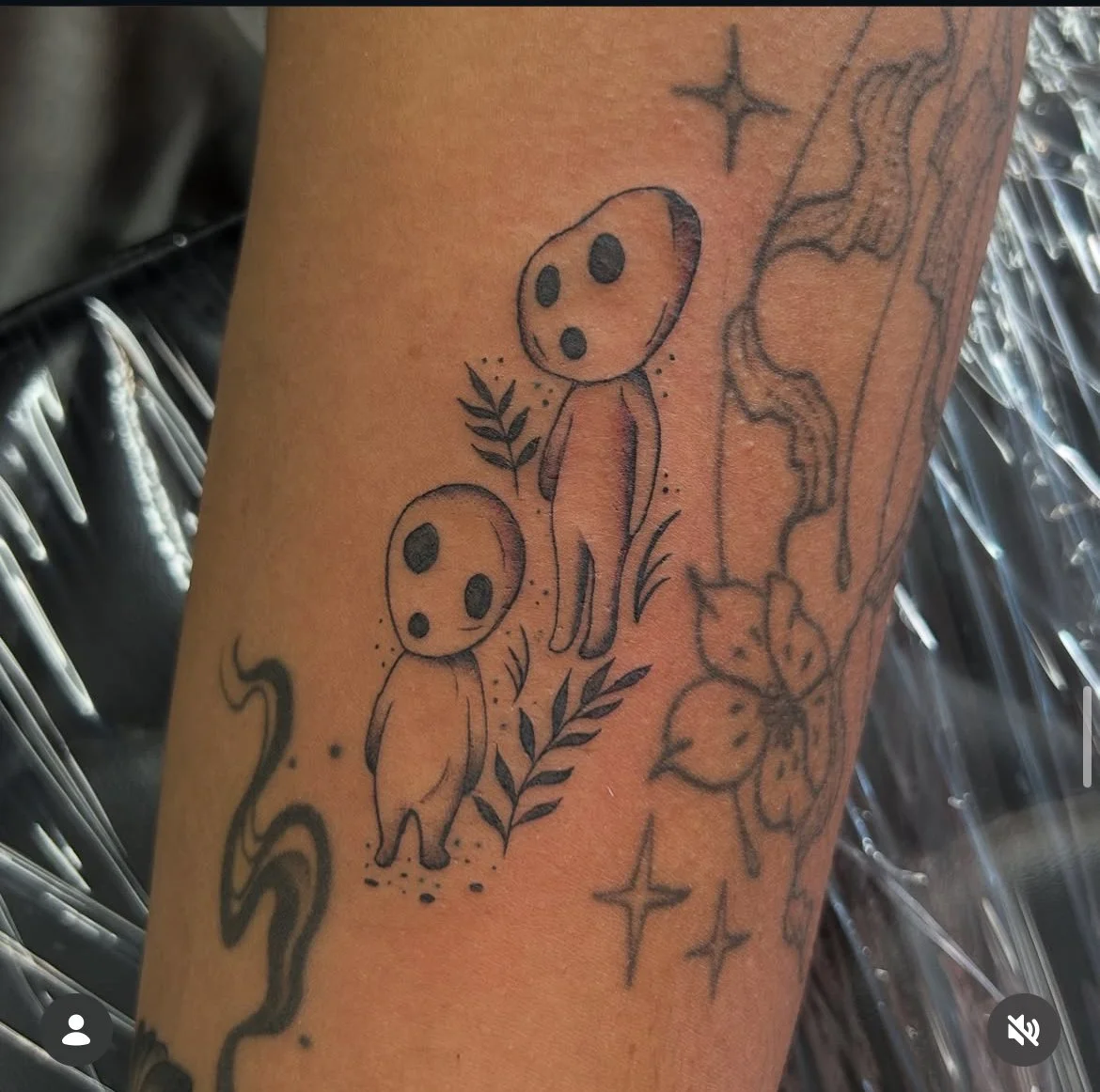 Tattoo of two cartoon-like figures with large round heads and black eyes, surrounded by plants and stars, on a person's arm. studio ghibli. kodama