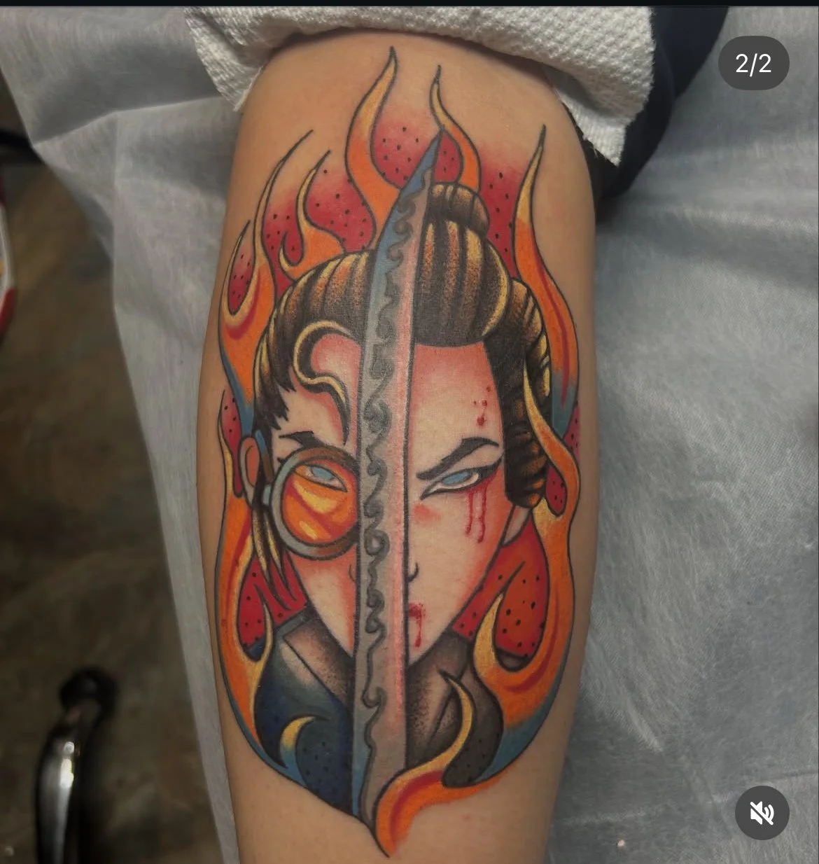 Tattoo of a woman's face behind a vertical sword, with flames around her, in a traditional tattoo style. blue eyed samuri.