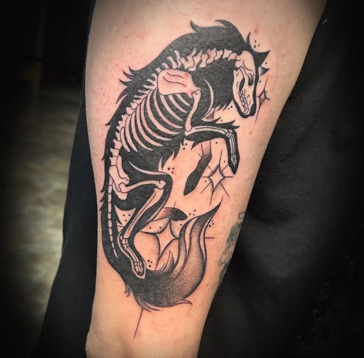 A black and gray tattoo of a horse skeleton with a mane, tail, and ribs, along with floral elements on skin.
