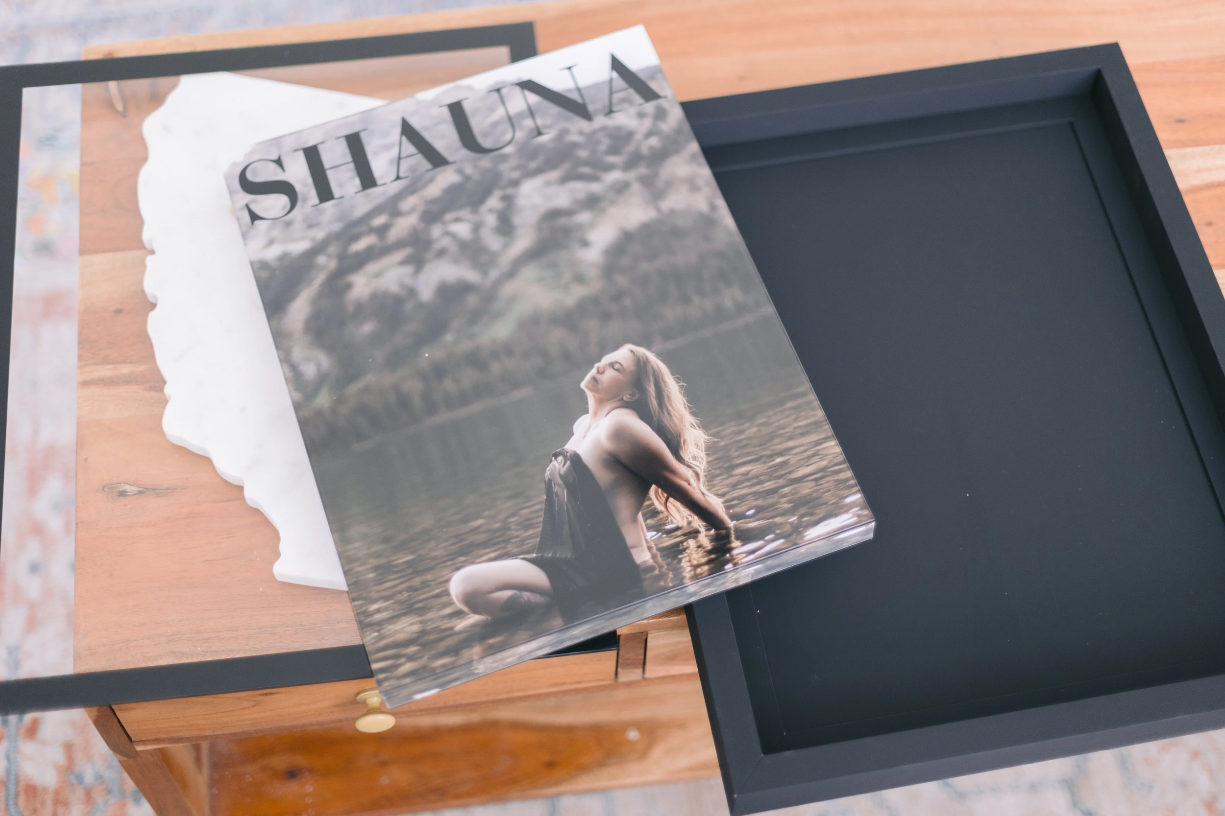 A photograph of a framed photograph of a woman sitting in water, with the word "SHANA" visibly printed on the photograph, placed on a wooden table with a black frame.