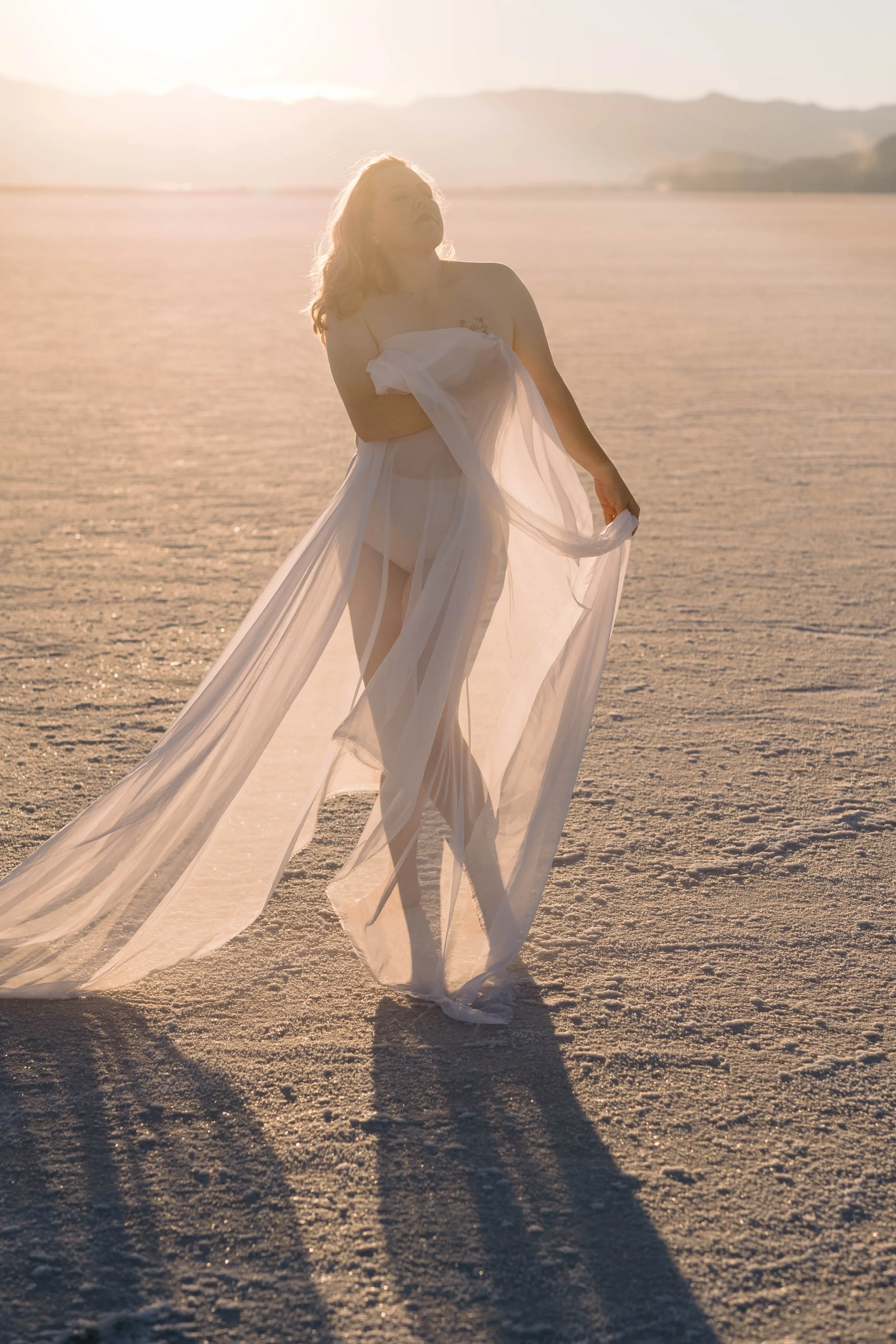 A woman standing in a desert at sunset, wearing a sheer white dress and holding part of it, with mountains in the background.
