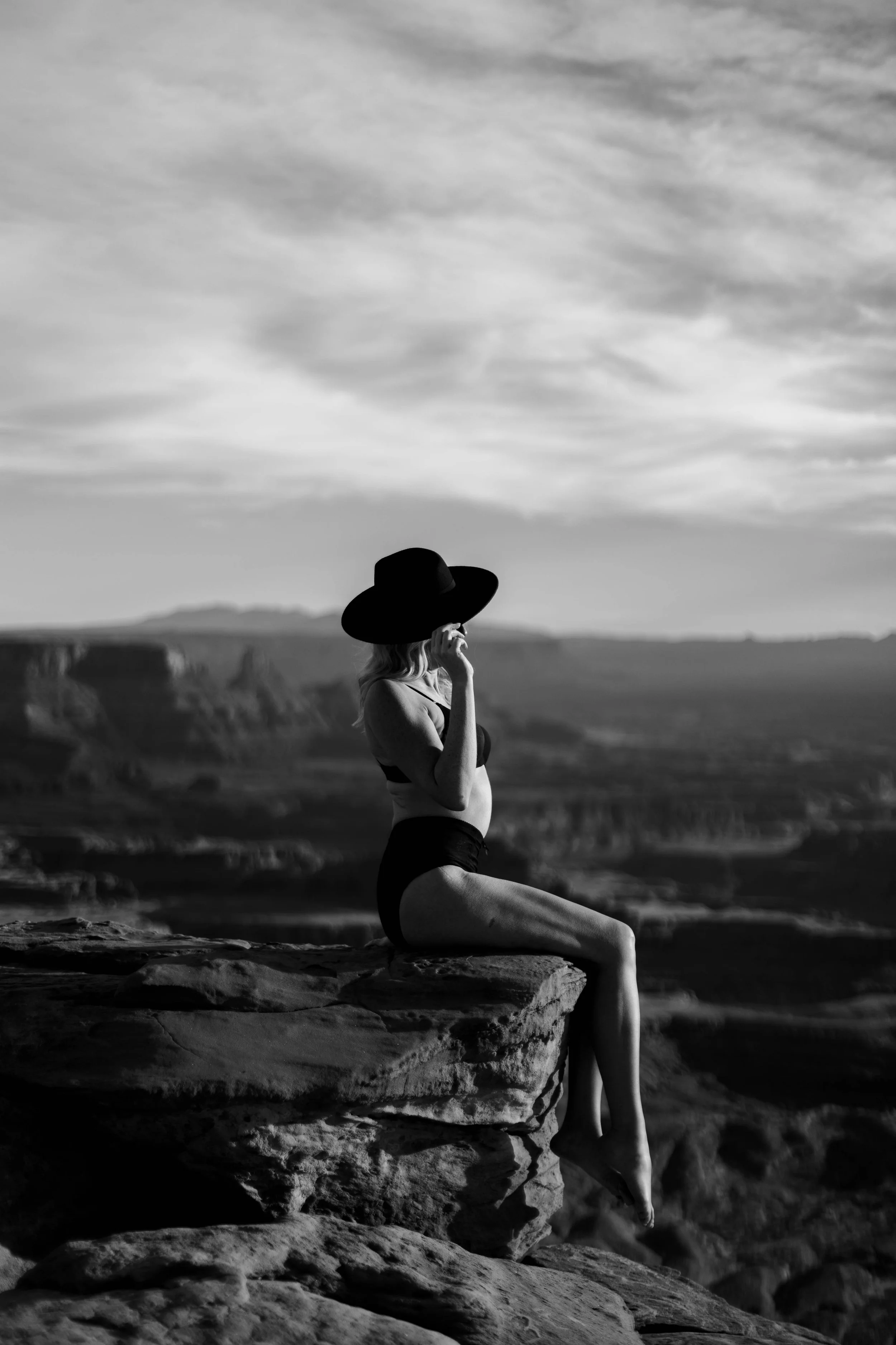 A woman in a black bikini and wide-brimmed hat sitting on a rock, with a vast landscape and cloudy sky in the background.