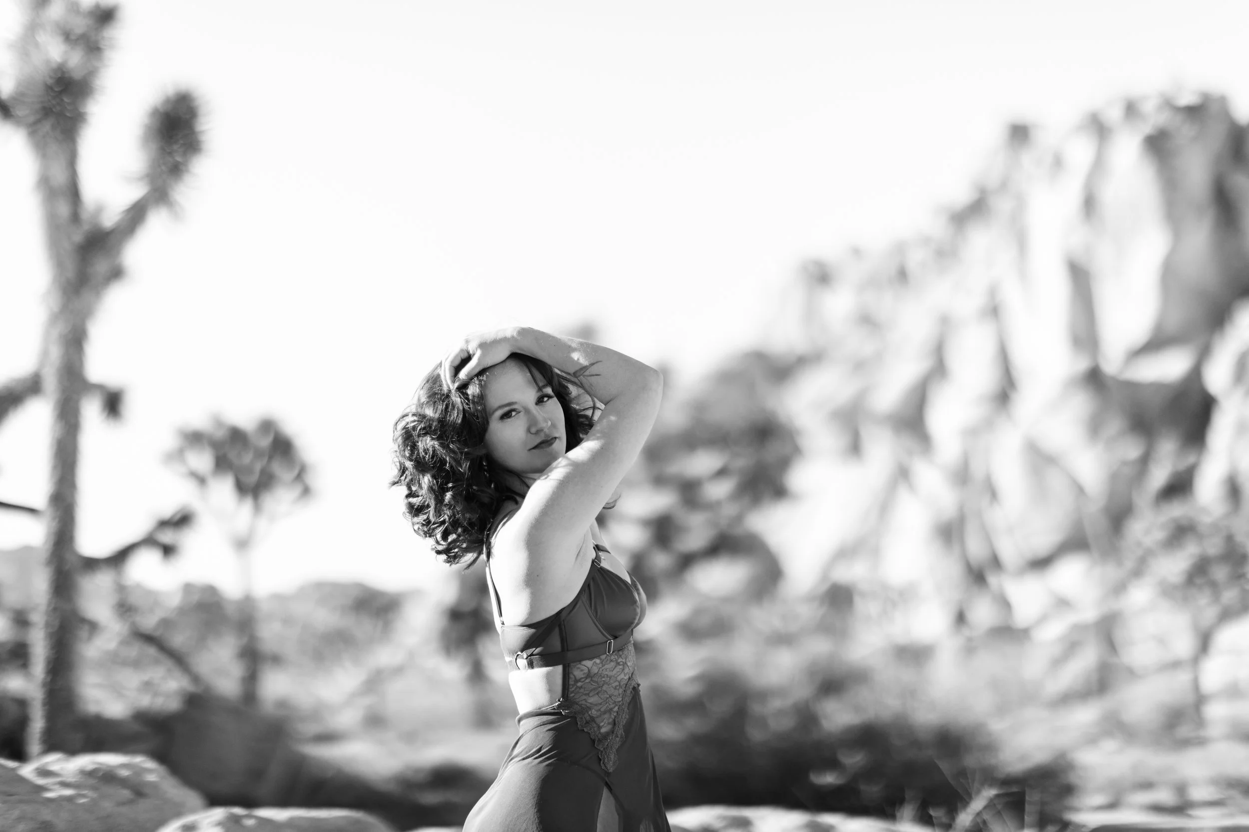 Black and white photo of a woman with curly hair outdoors near rocks and trees, posing with one hand on her head.