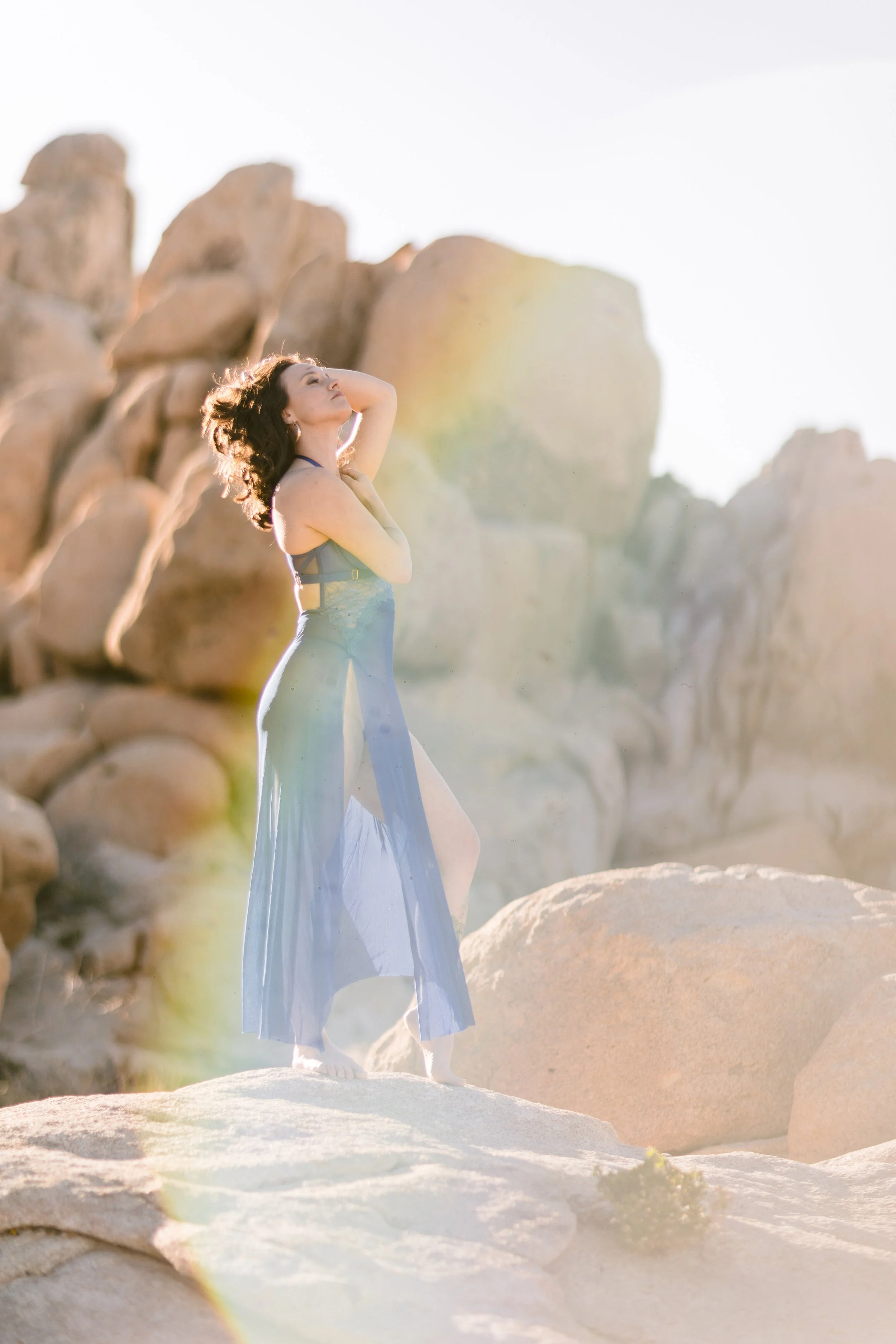 Woman in a flowing dress posing among large rocks in a desert landscape with bright sunlight.