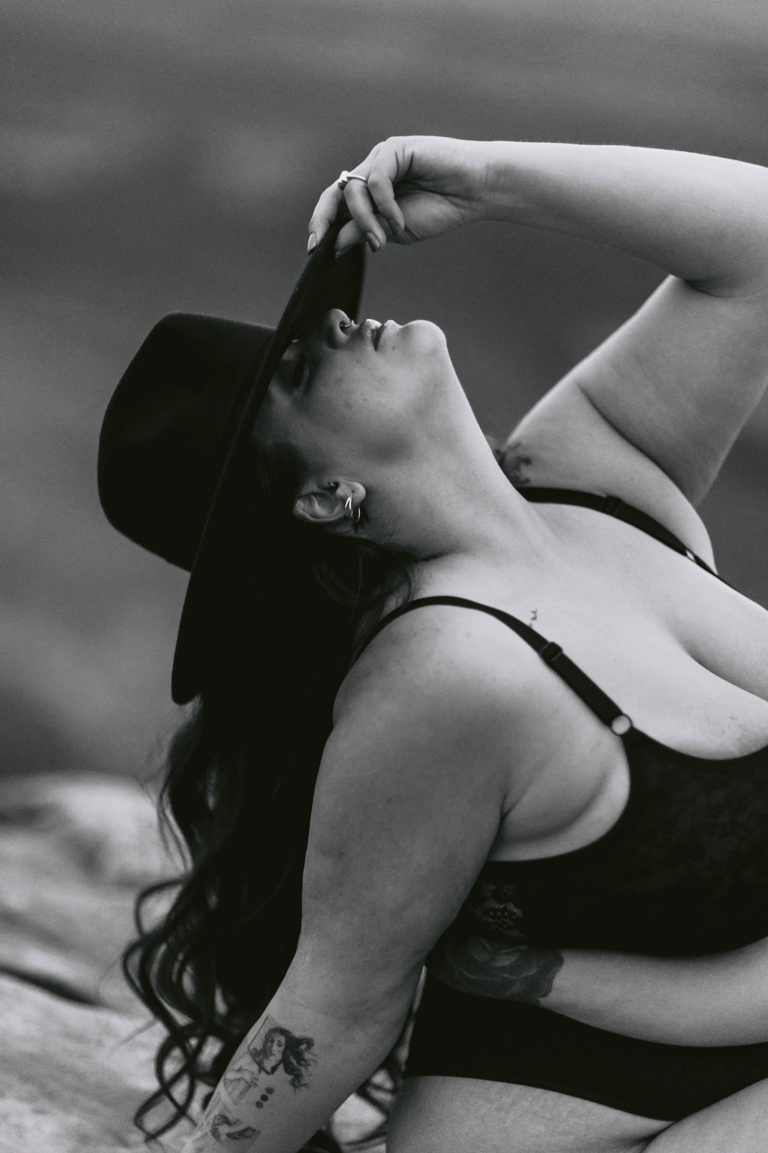 A woman with long wavy hair, wearing a hat and a black bra, lying on the floor. She is holding the brim of her hat and looking up. She has a tattoo of a vintage woman on her forearm and earrings.