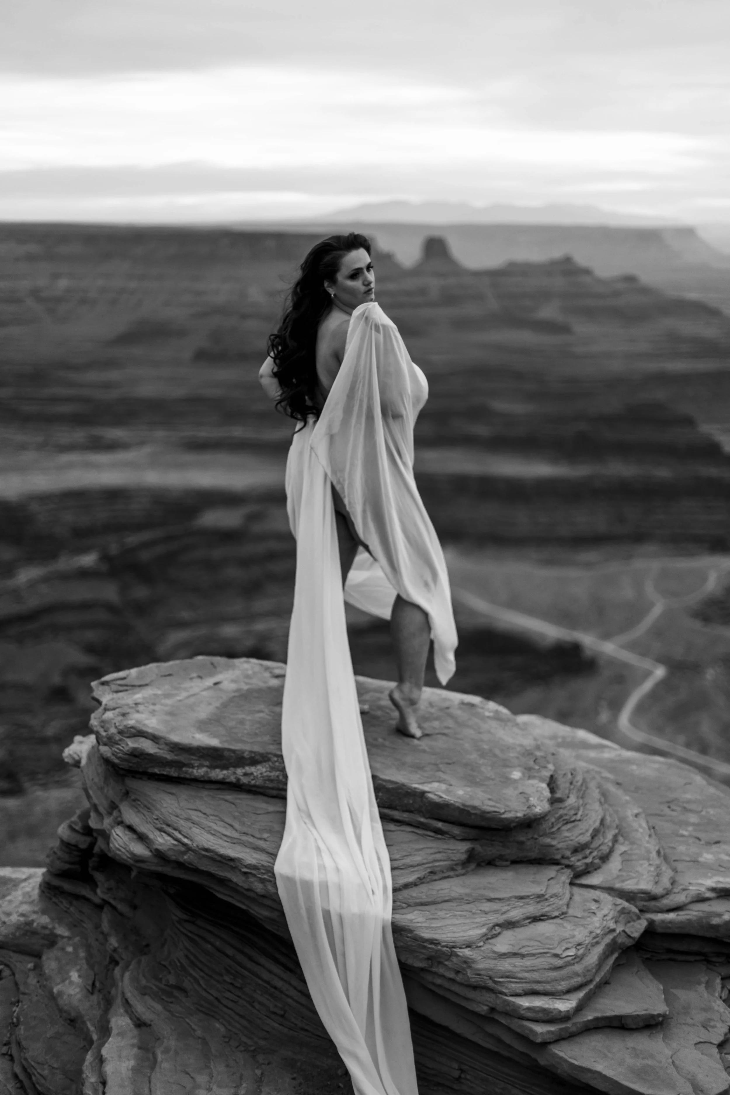 A woman in a flowing white dress stands on a large rock formation in a desert landscape, with mesas and plateaus visible in the background, in black and white.