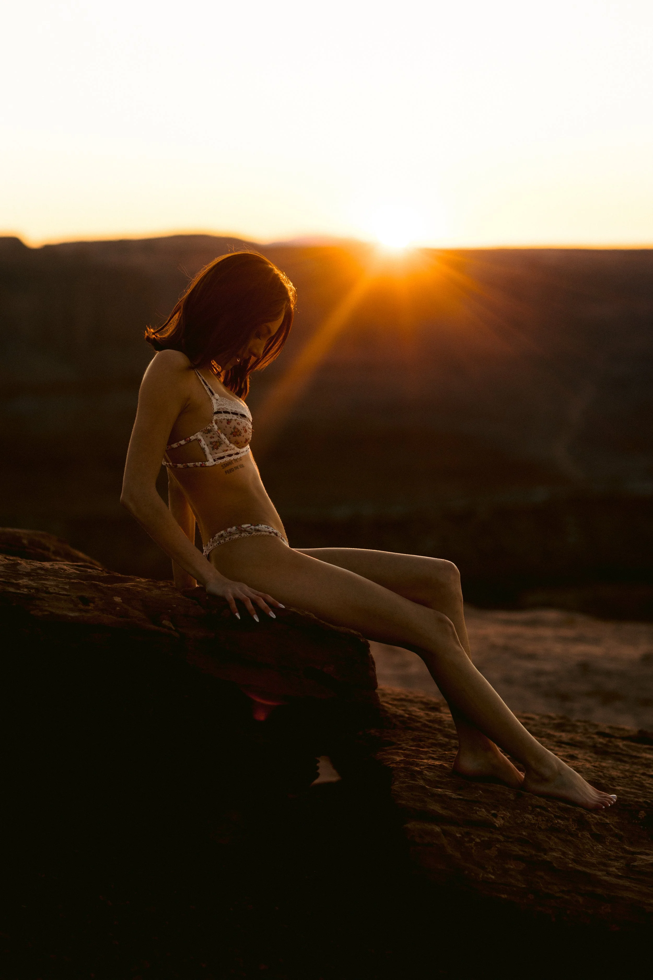 A woman with red hair sitting on a rock at sunset, wearing floral lingerie, with the sun setting behind her in a desert landscape.