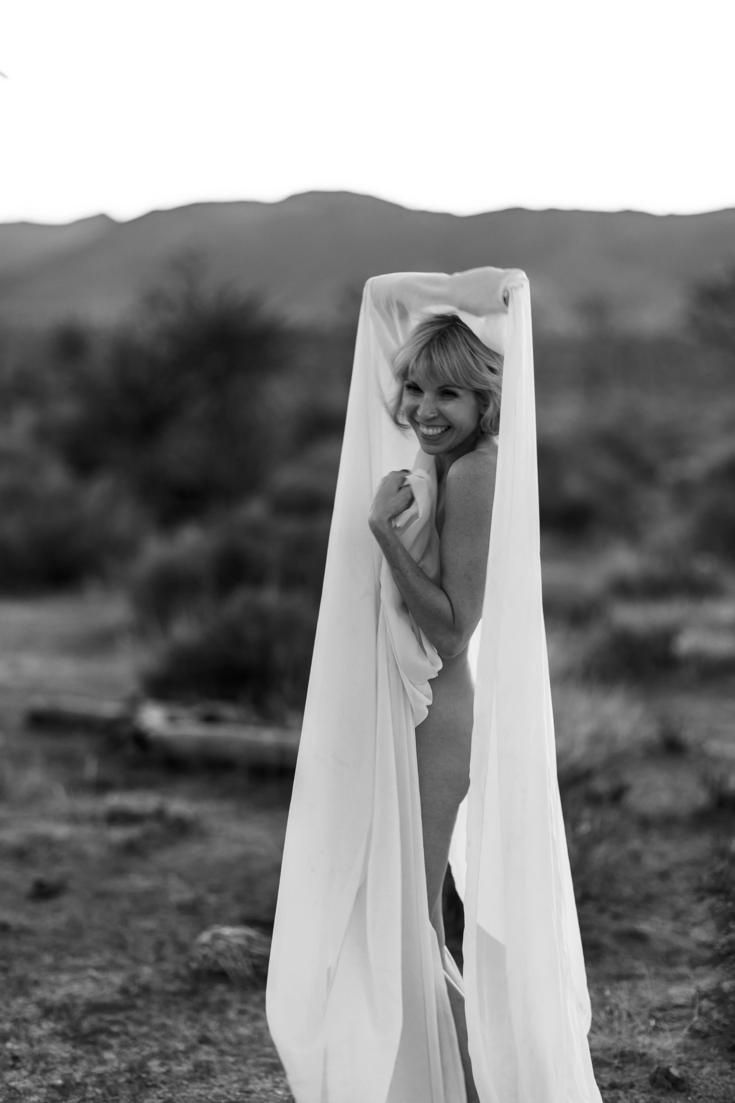 A woman smiling and covering her face with a white cloth in an outdoor natural landscape.