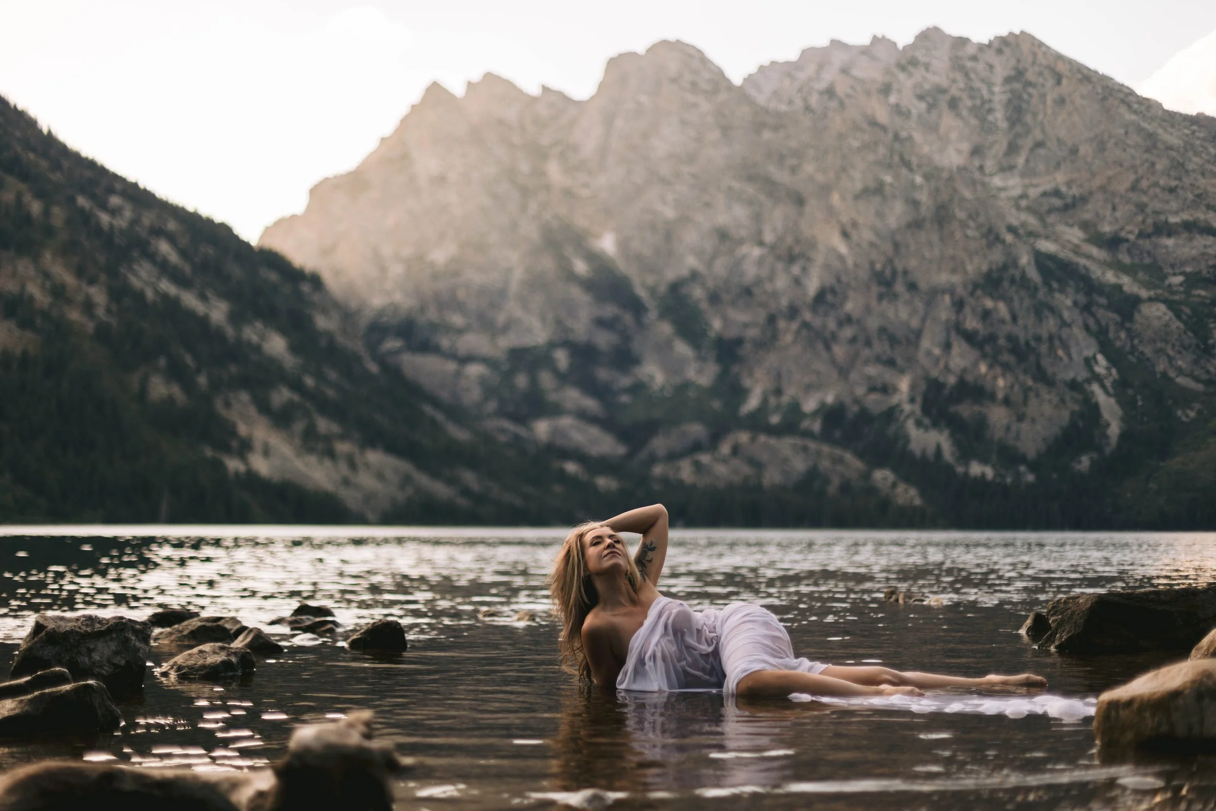 A woman in a white dress is lying partially in the water at the edge of a lake with mountains in the background.