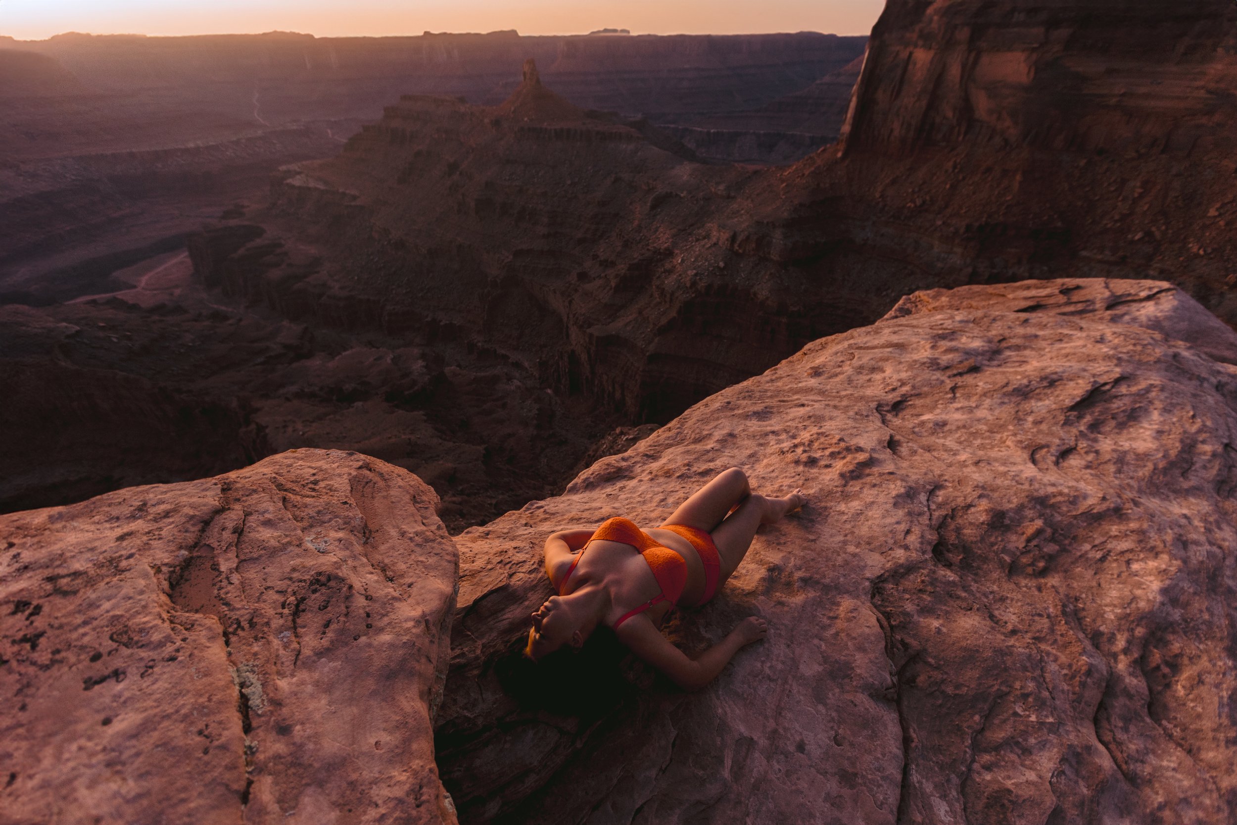 Person lying on a rocky ledge overlooking the Grand Canyon during sunset, wearing an orange swimsuit.