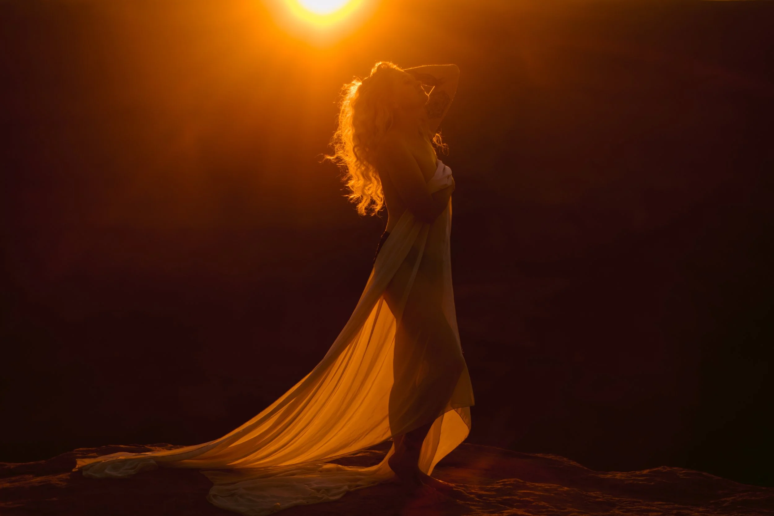 A woman with long curly hair standing in a dark space, illuminated by a bright orange-yellow light from above. She wears flowing, layered clothing and is holding her head with one hand, creating a silhouette with a warm glow.