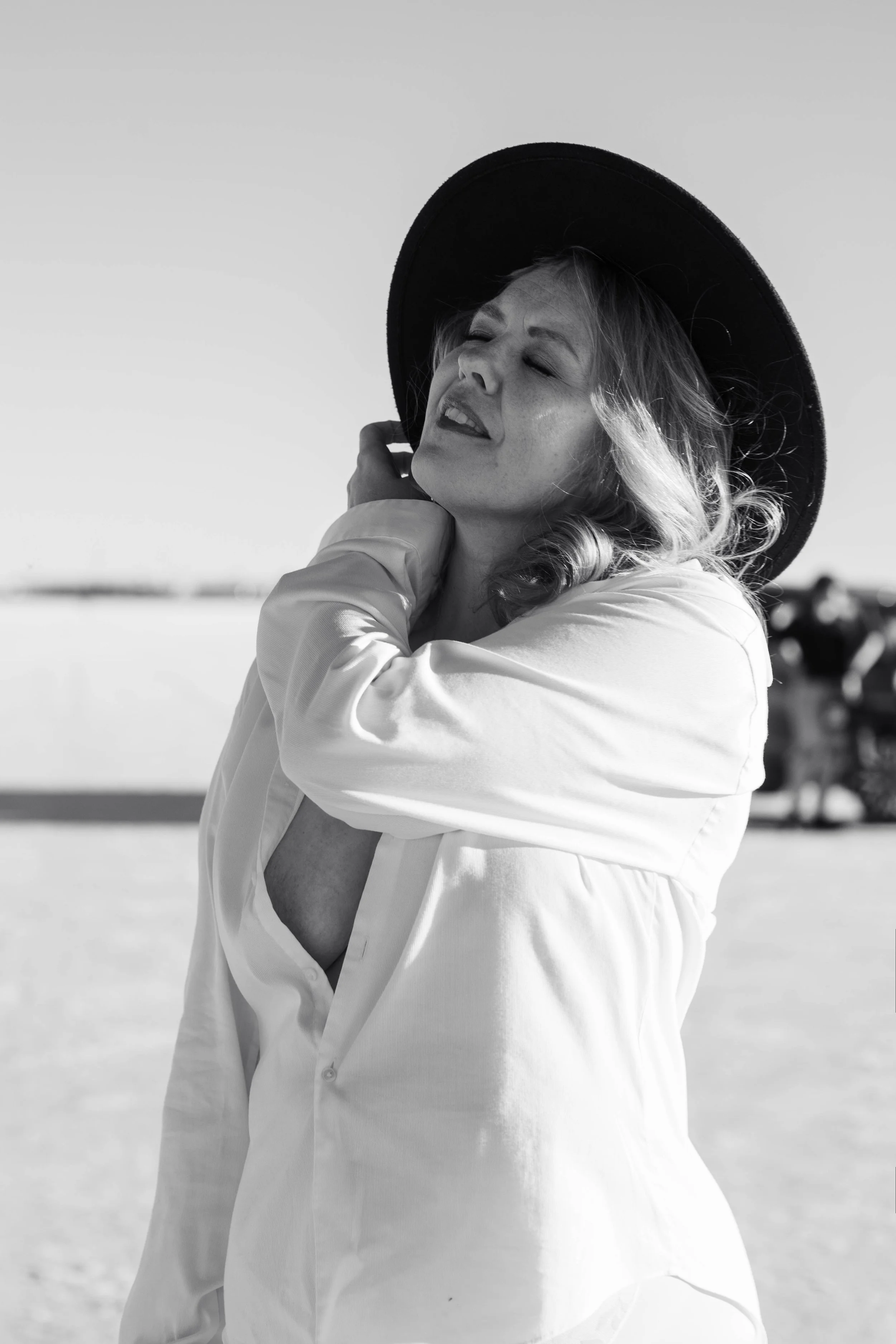 A woman with wavy hair and a wide-brimmed hat, wearing a light-colored blouse, sitting or standing outdoors with sunlight, her eyes closed, and her hand near her neck.