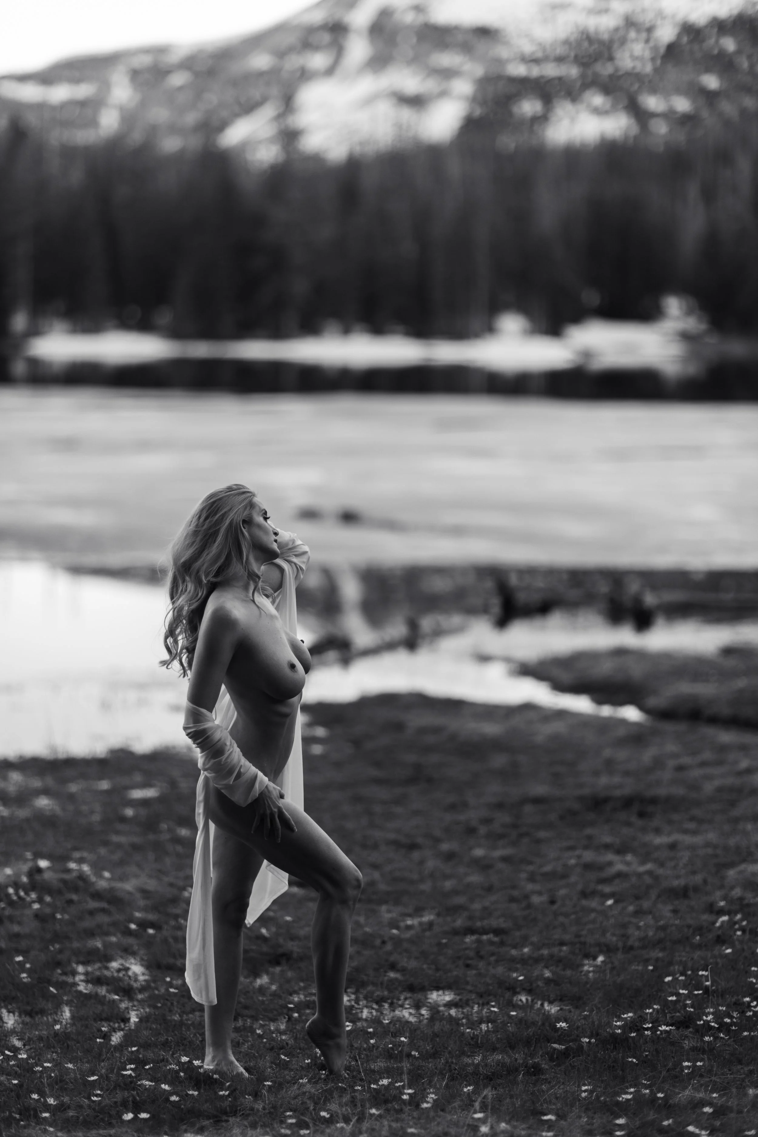 A black and white photo of a woman with long hair standing outdoors near a body of water. She is topless, wearing a white skirt, and has her right hand on her hip, gazing to her right. The background shows a hilly or mountainous landscape with trees.
