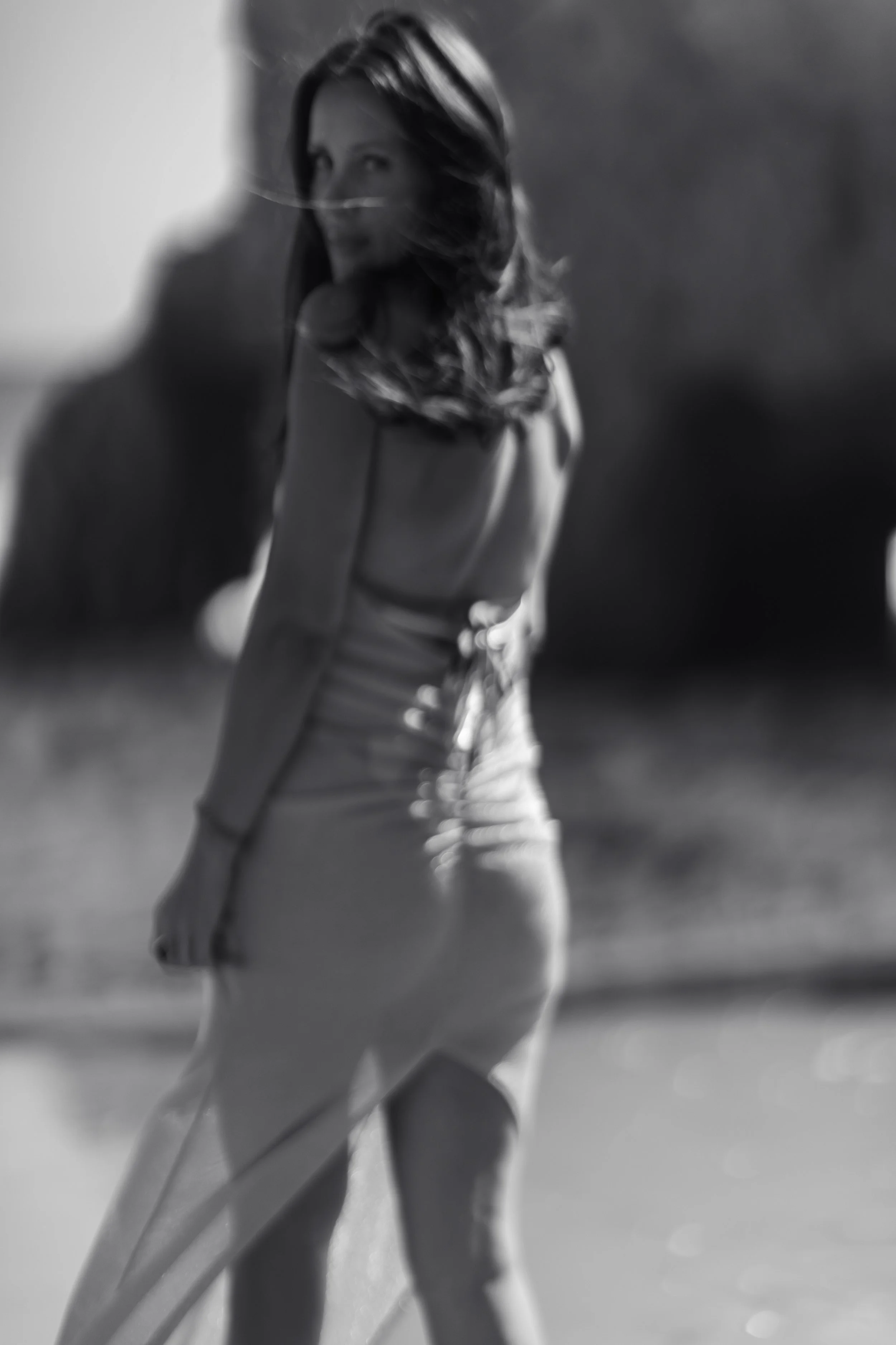 Black and white photo of a woman standing in a desert, with her back to the camera, wearing a sleeveless top and pants, holding a surfboard, with windswept hair and a distant gaze.