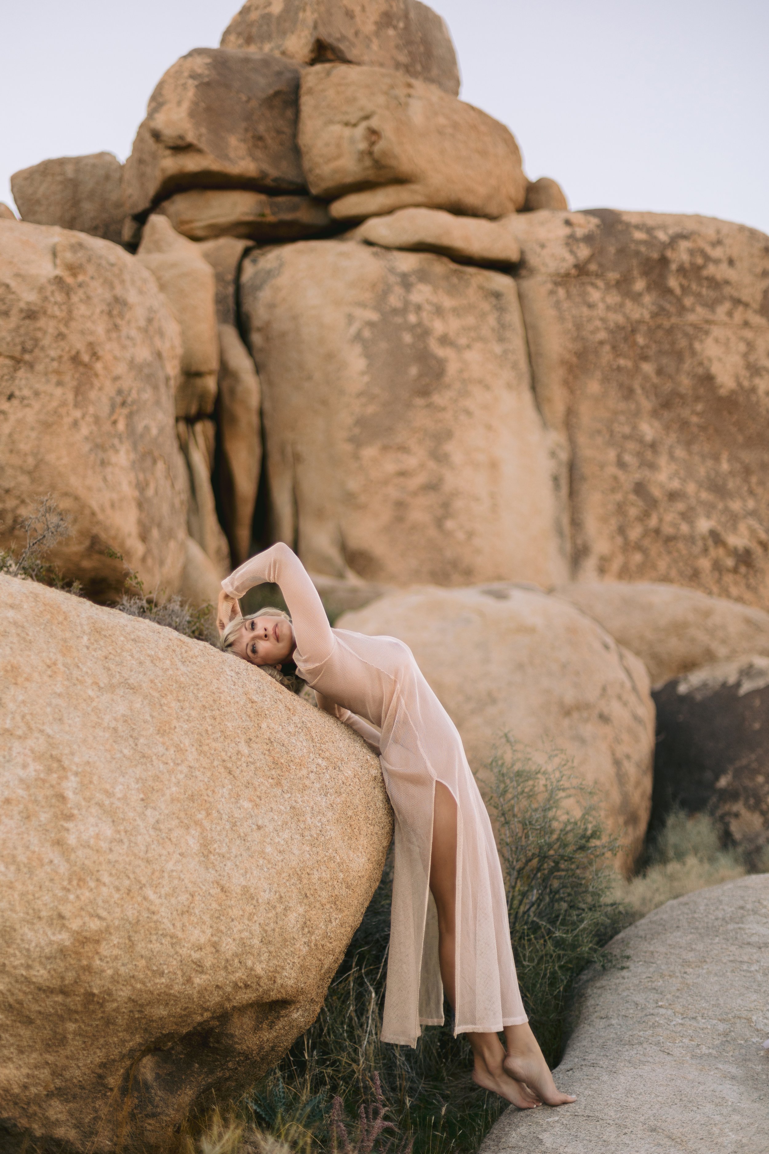 A woman with blonde hair in a beige dress lies on a large rock, with her head tilted and her hand on her forehead, surrounded by a desert landscape with large rocks and sparse vegetation.