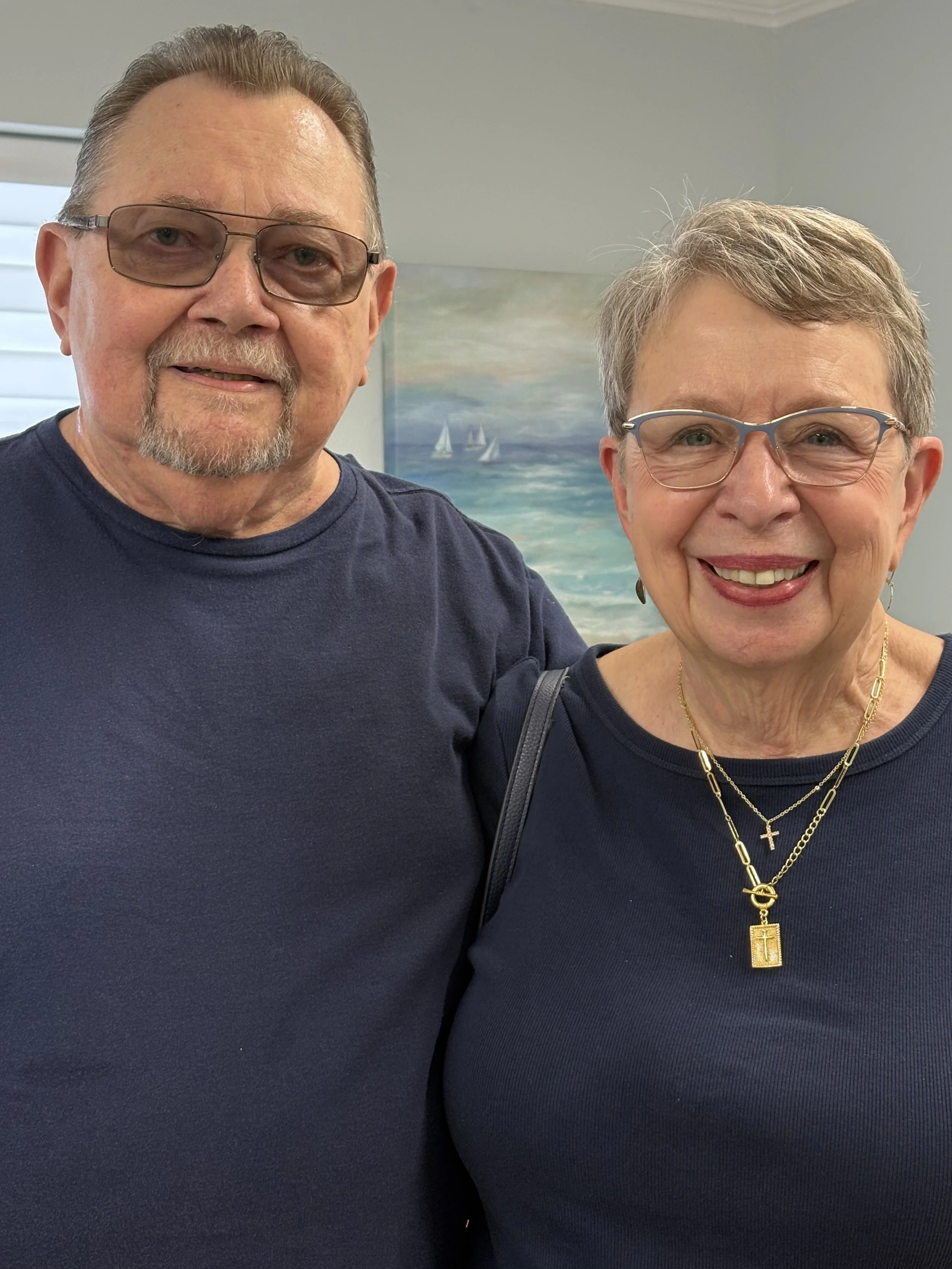 An elderly man and woman sharing a joyful moment, with the man leaning forward and smiling at the woman, who is also smiling and looking at him. They are indoors with a dark background.