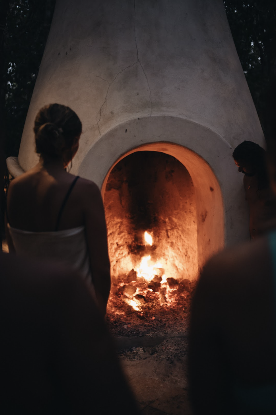People gathered around a glowing outdoor clay fire oven at dusk.