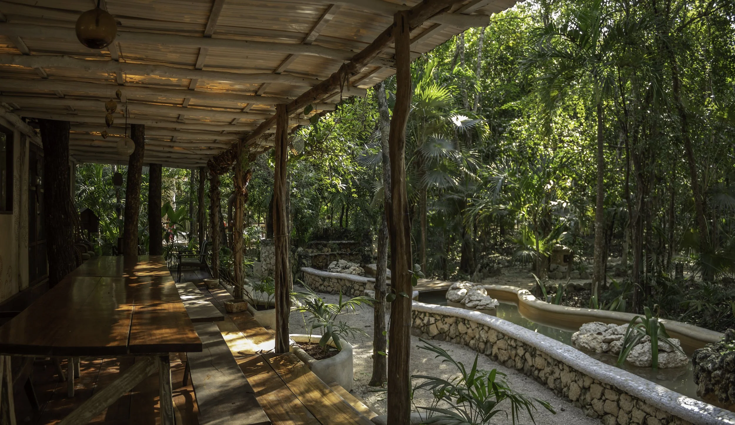 Rustic wooden patio with a long table beside a stone-lined water feature in a lush tropical jungle garden