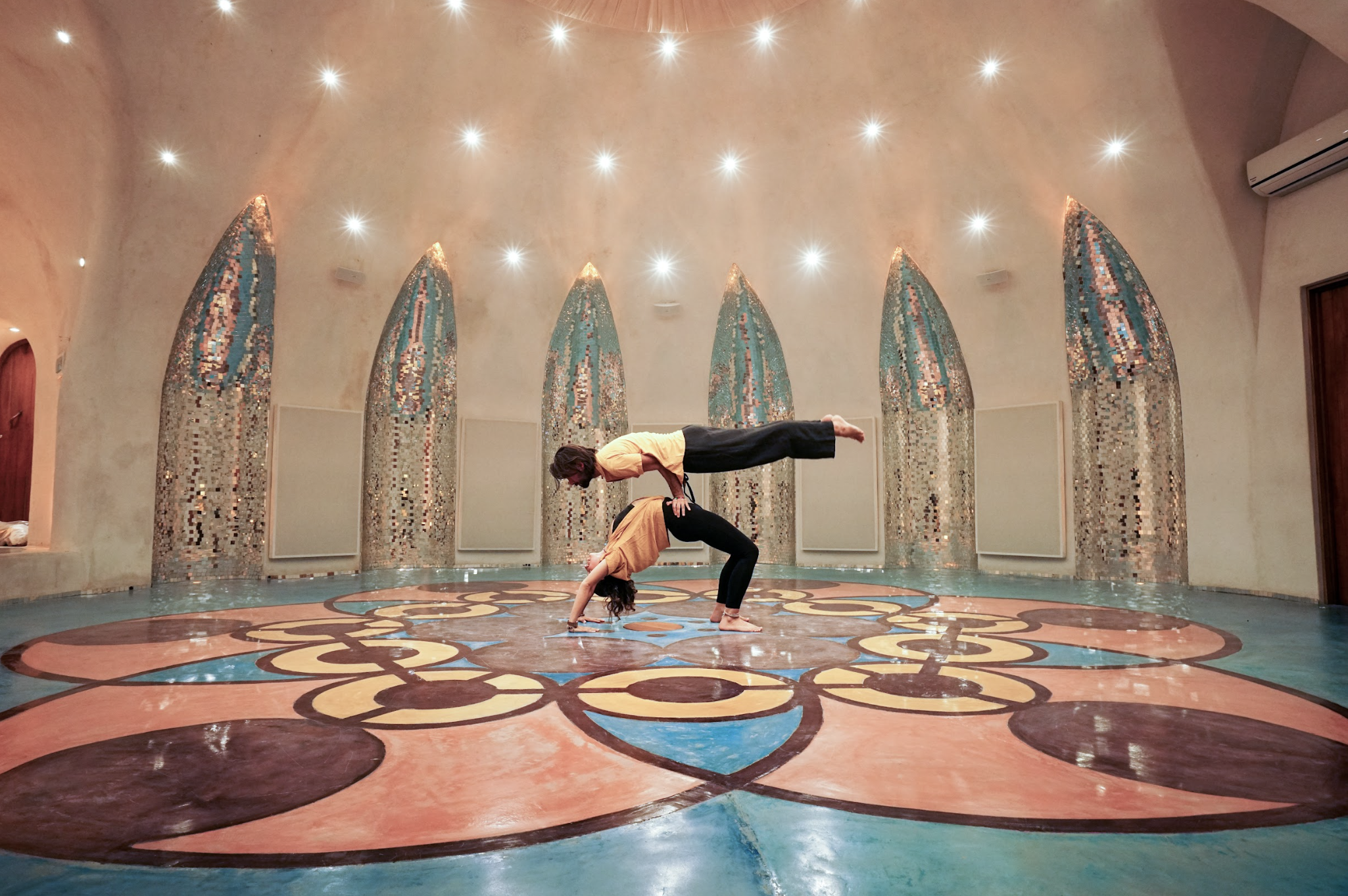 Two people performing an acro yoga pose on a colorful mosaic floor in an ornate indoor room.