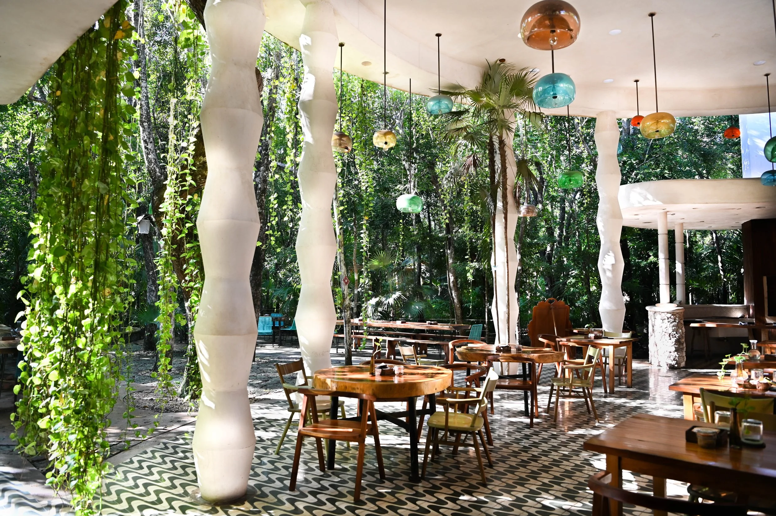 Open-air jungle restaurant with white sculptural columns, colorful pendant lights, wooden tables, and lush tropical greenery