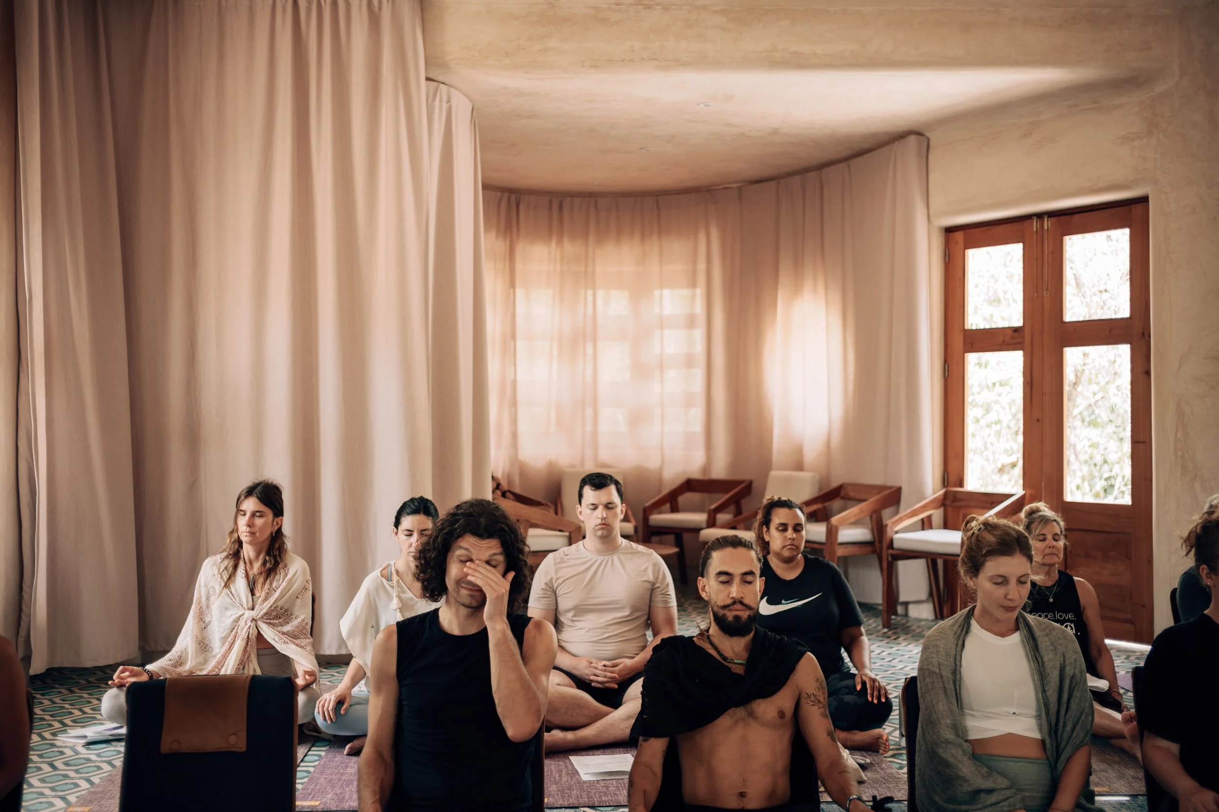 Group meditating in a sunlit room with sheer curtains and wooden windows