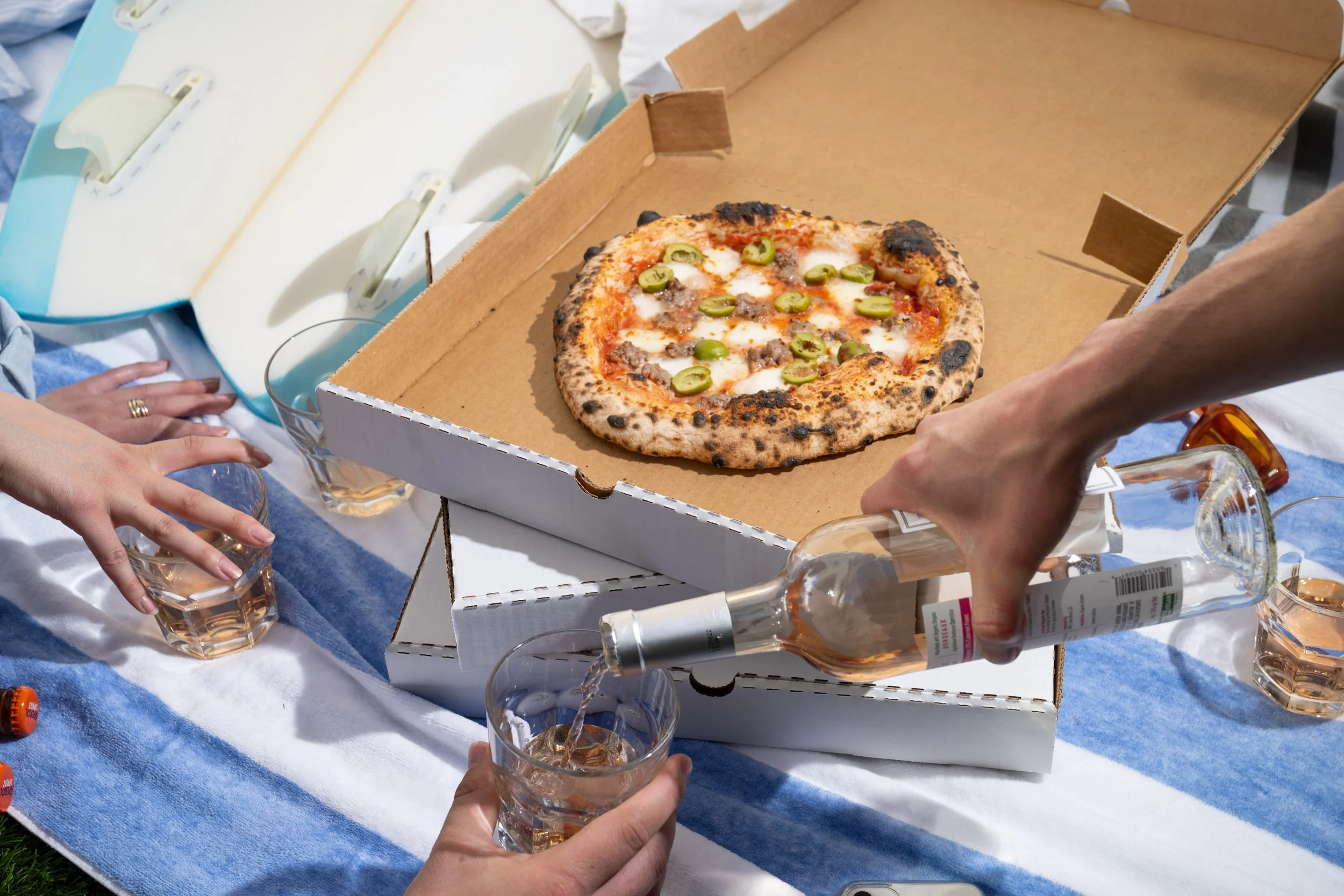 People celebrating outdoors with pizza and drinks on a table covered with a blue and white tablecloth. A hand is pouring rosé wine into a glass, and there are glasses of wine and bottles on the table.