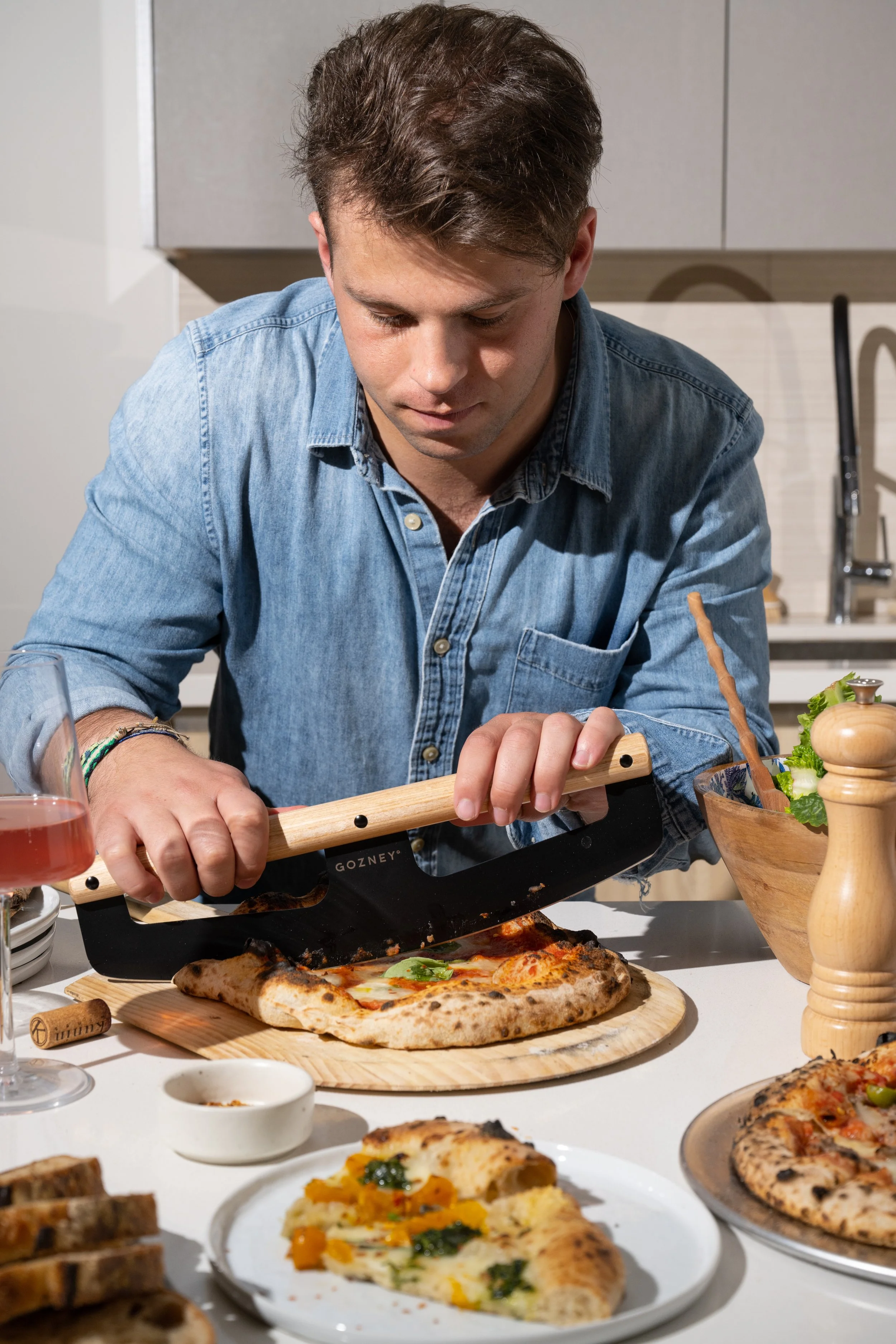A young man in a denim shirt is slicing a freshly baked pizza with a sharp knife in a modern kitchen.
