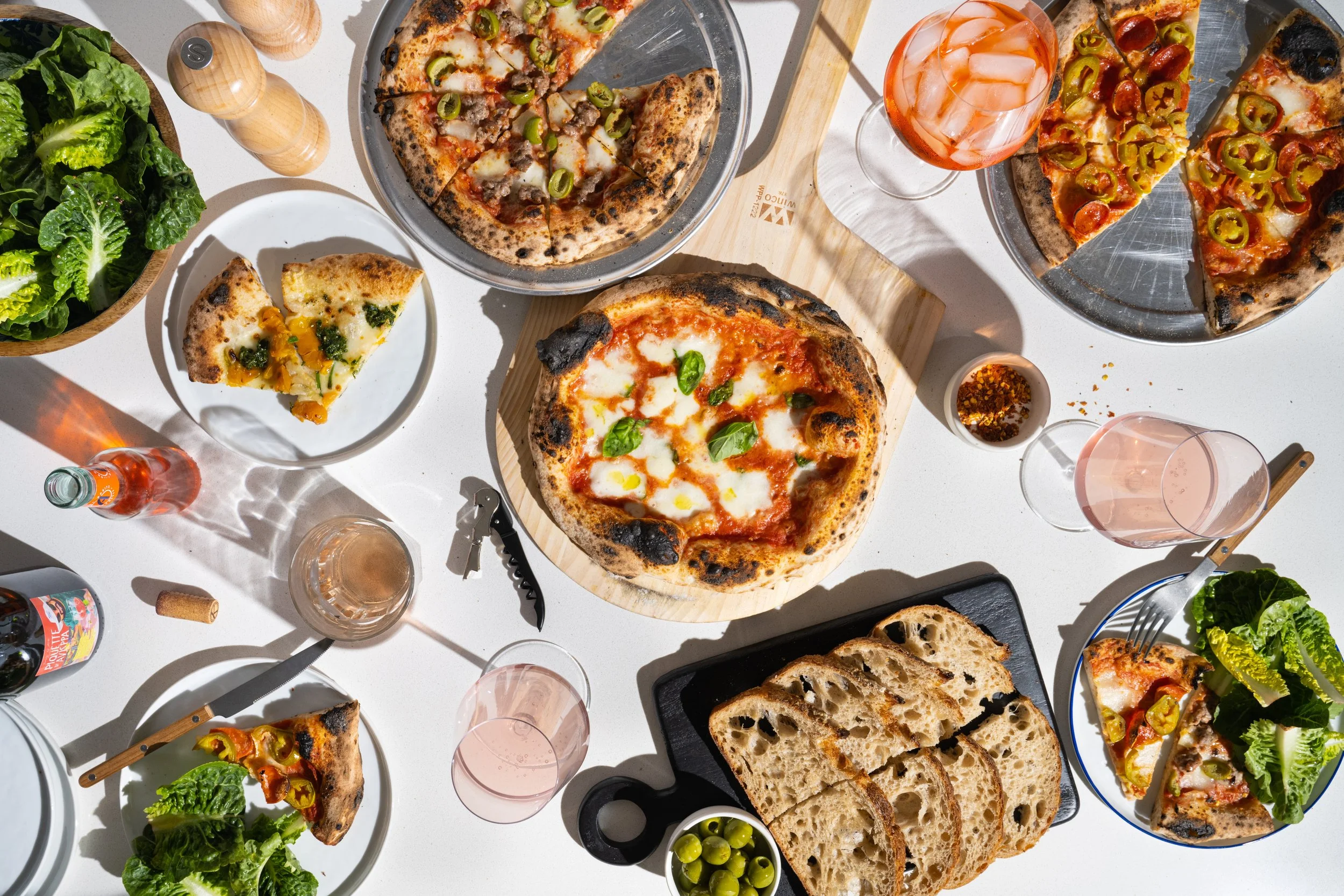 Assorted pizzas with toppings like cheese, pepperoni, sausage, green peppers, and basil, served on a white table alongside a bowl of green grapes, slices of bread, a salad with lettuce, and several glasses of pink-colored drinks.