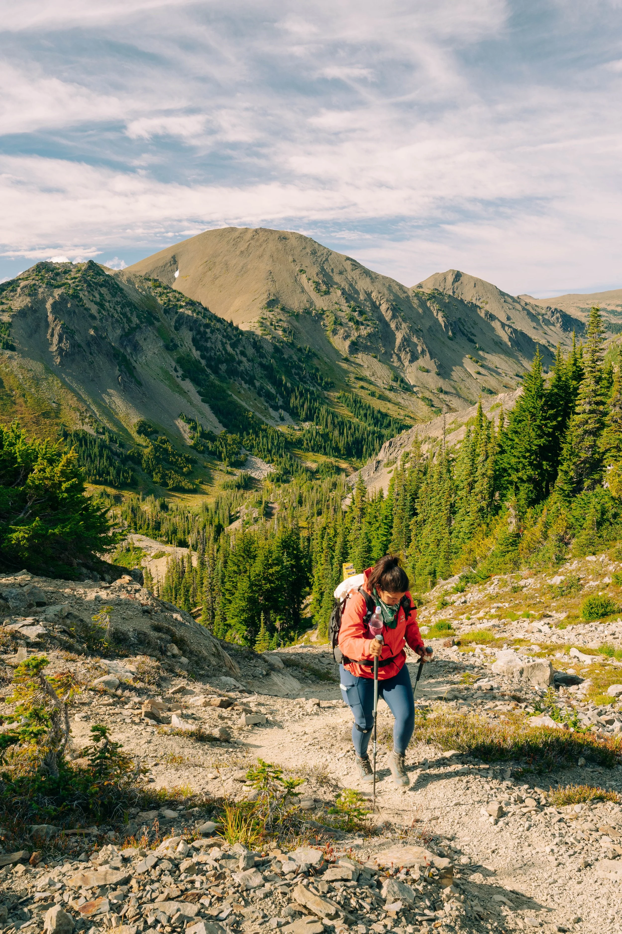 A woman hiking on a rocky mountain trail surrounded by green trees and mountains in the distance.