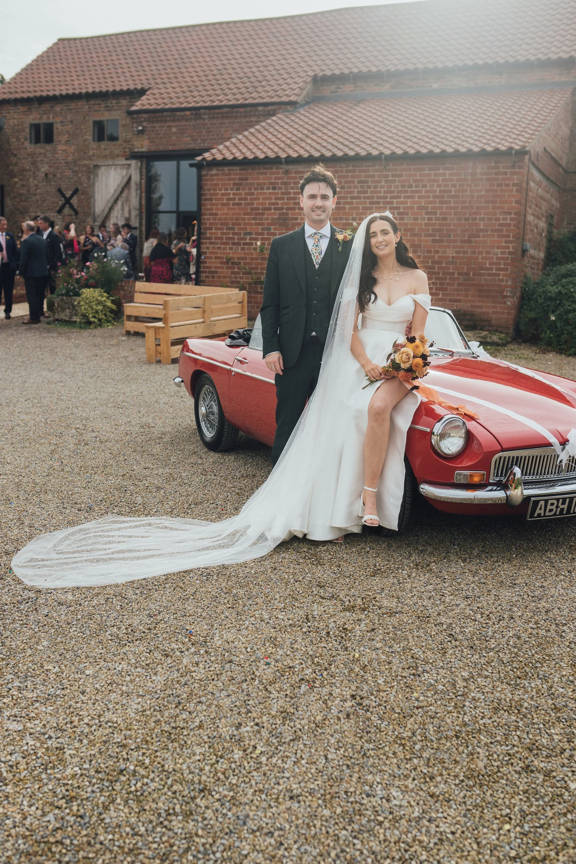Bride and groom standing next to a red classic car at their wedding celebration, with guests in the background outside a brick building.