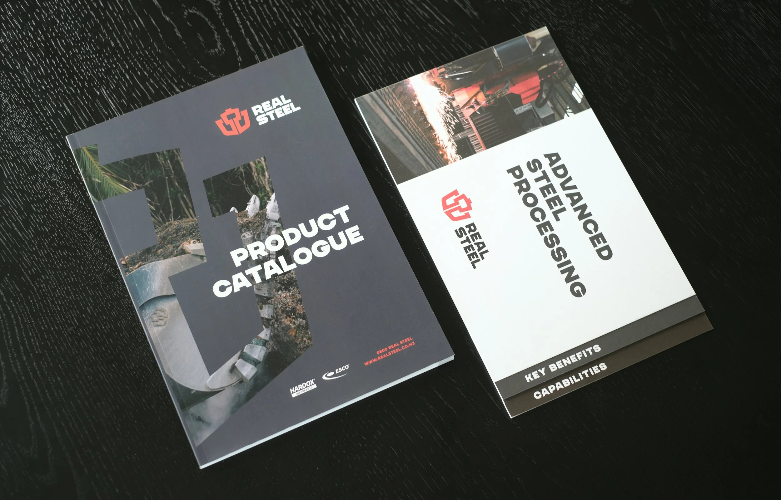 A Real Steel Product Catalogue book next to a document called "Advanced Streel Processing", both laid out on a black wood table.