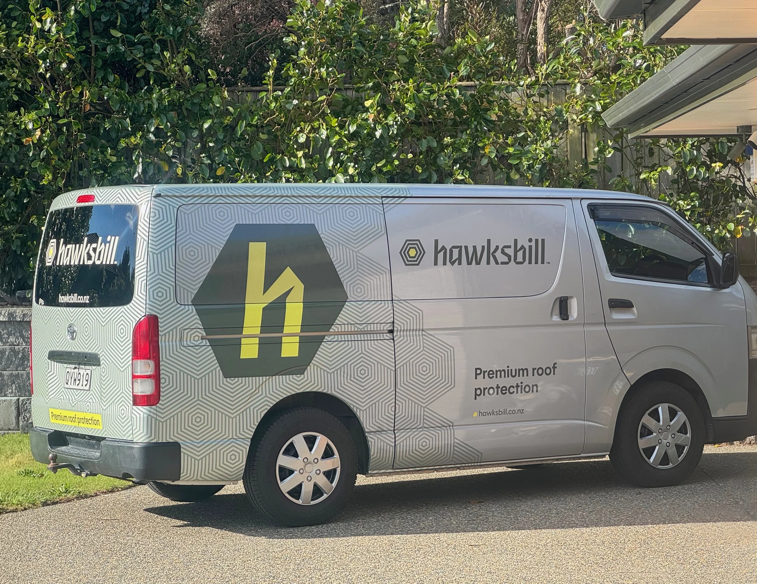 A silver van parked outside a house with branded vehicle graphics – a large green hexagon with an 'h' in the middle, a hexagon pattern covering the back and sides with the Hawksbill logo on the side and back window.
