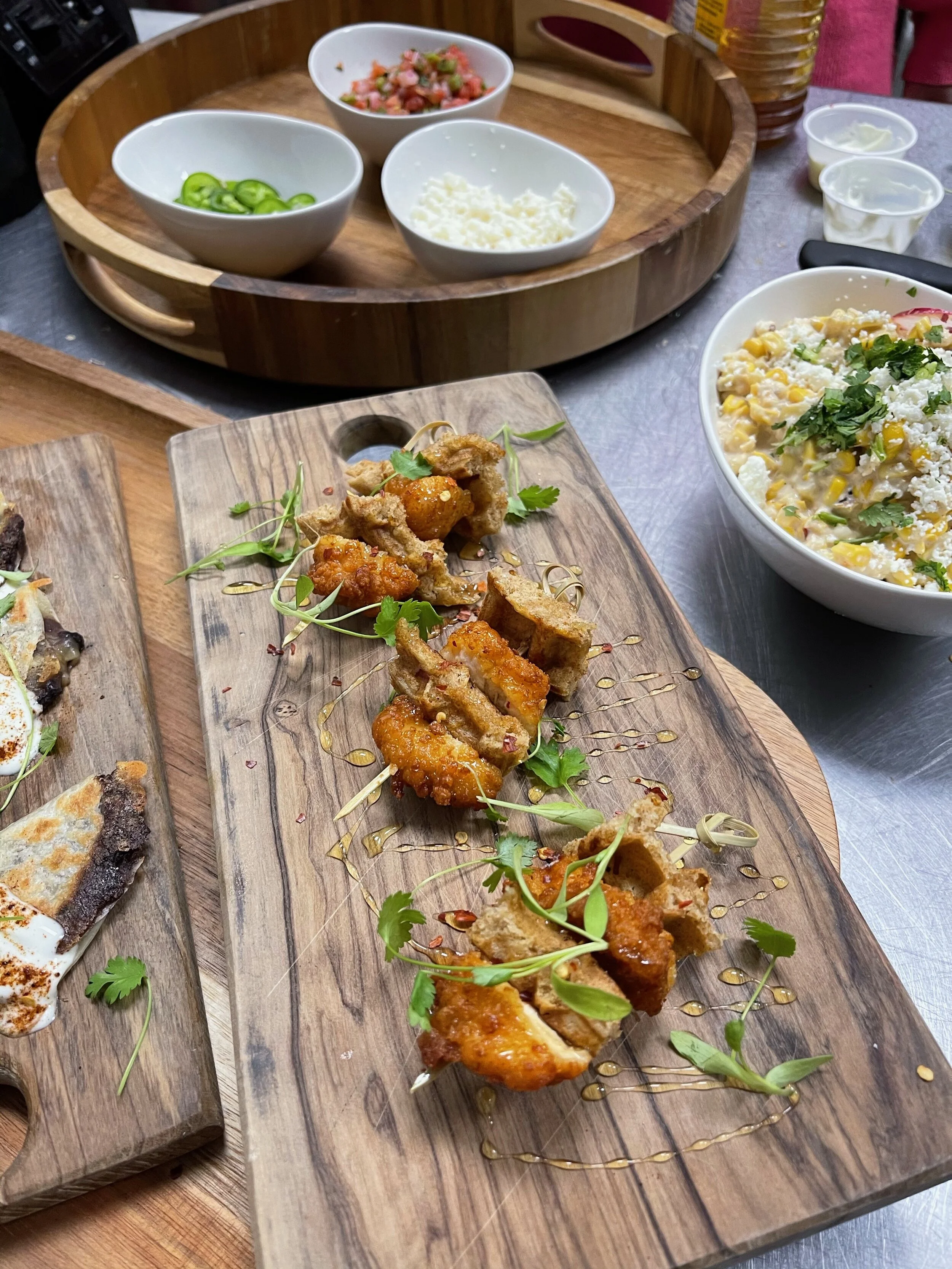 Assorted Mexican food setup with garnishes including fried appetizers on wooden boards, bowls of sliced jalapeños, grated cheese, and salsa, alongside a bowl of corn salad.