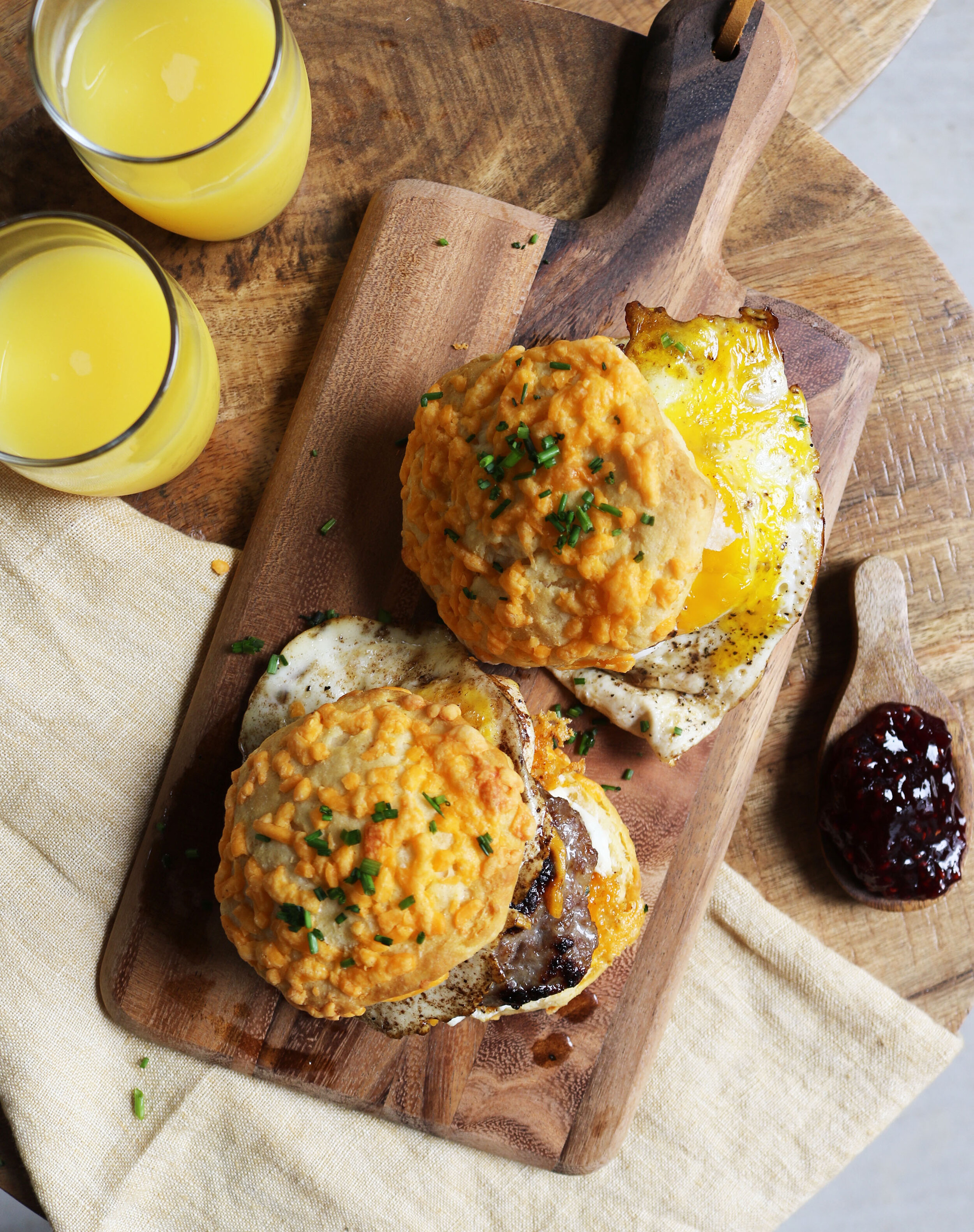 Breakfast sandwiches with biscuits, fried eggs, and sausage on a wooden board, accompanied by jelly and glasses of orange juice.