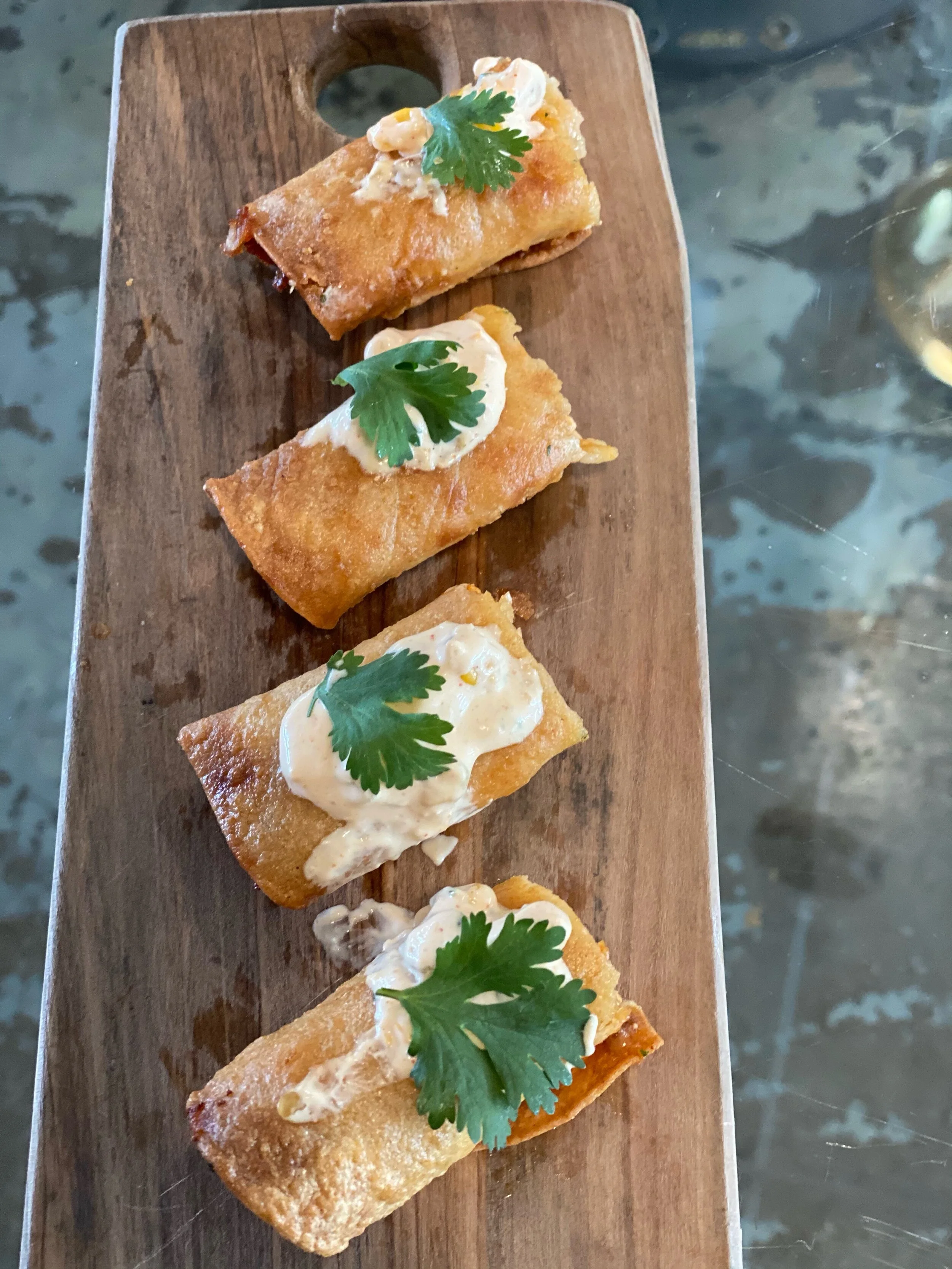Four crispy rolled appetizers on a wooden serving board, topped with creamy sauce and cilantro.