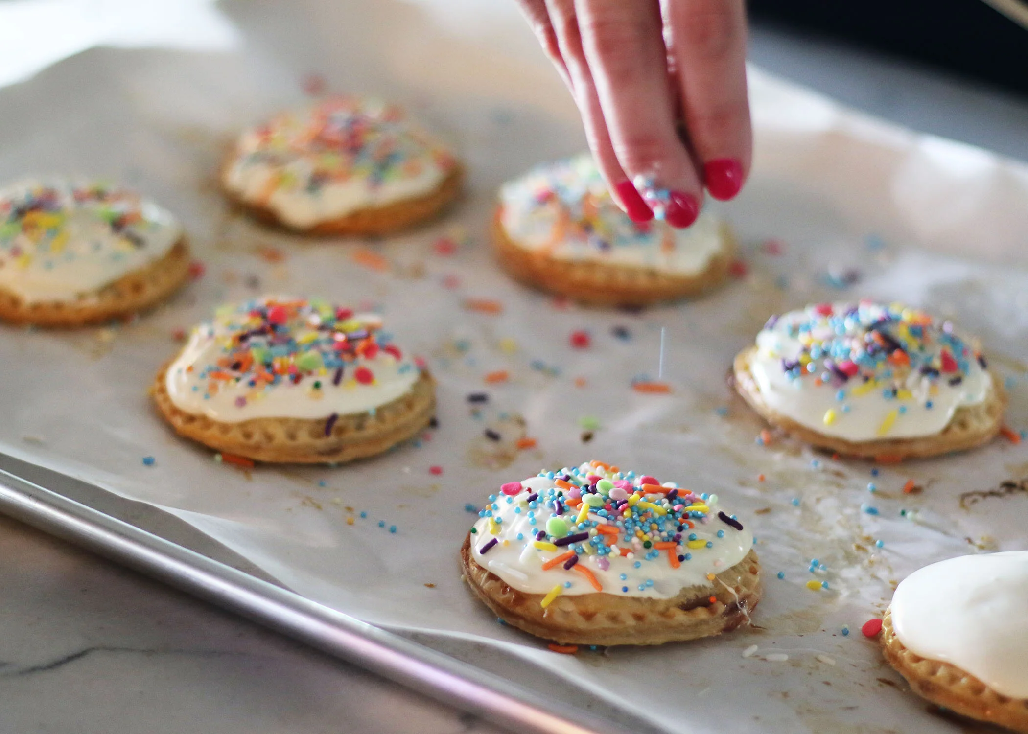 Hand sprinkling colorful sprinkles on frosted cookies on a baking sheet.