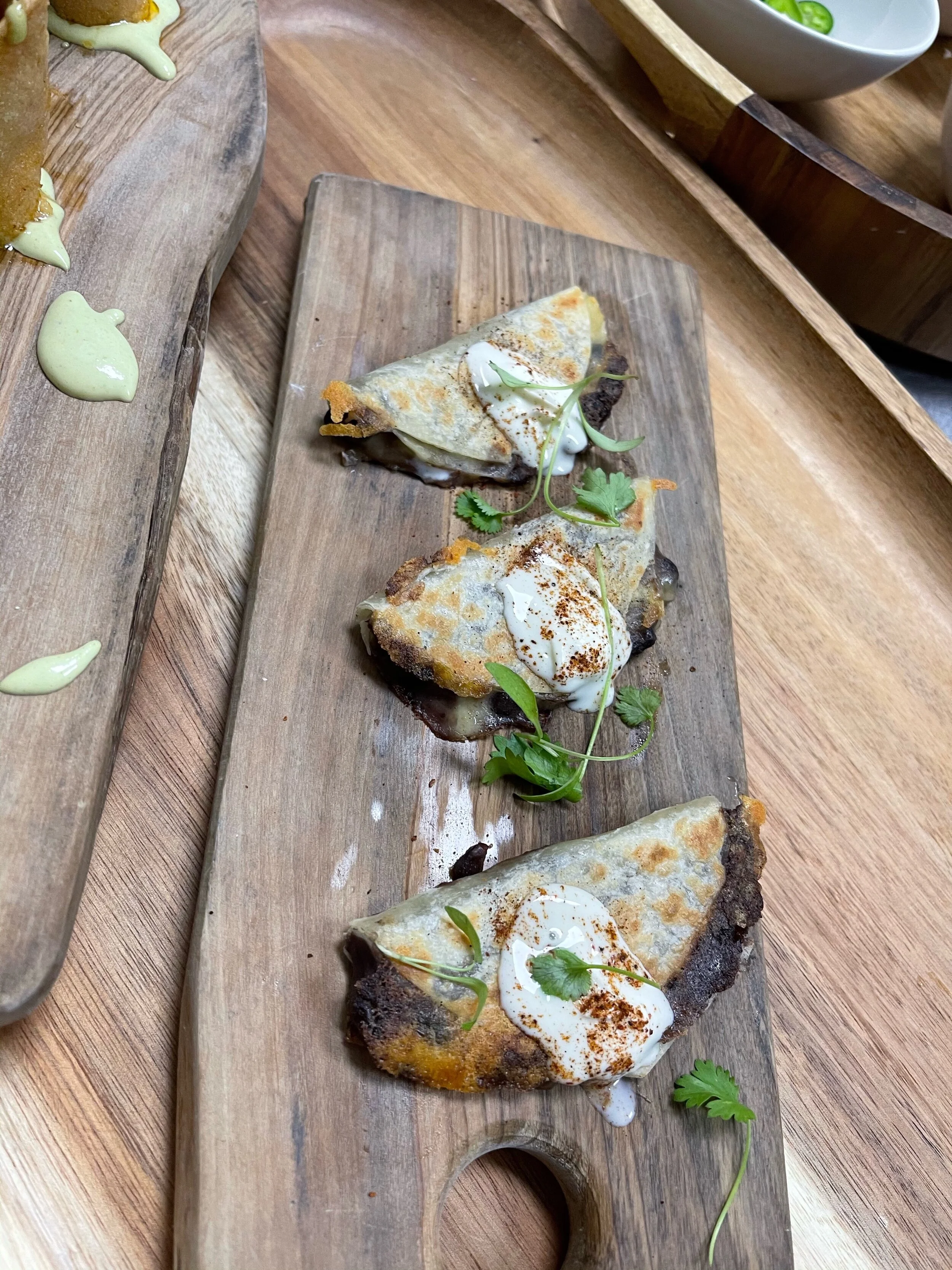 Three small folded tacos with sour cream, cilantro, and spices on a wooden serving board.